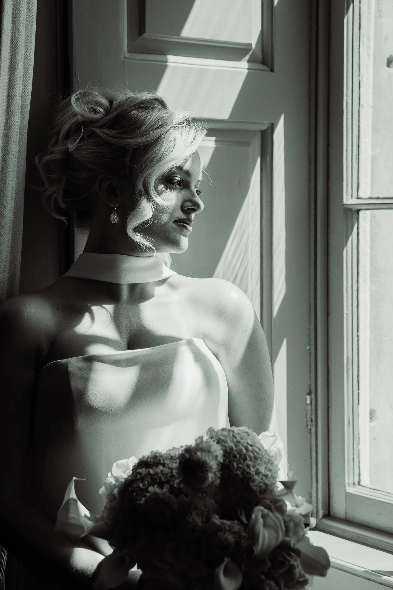 A woman with styled blonde hair in an elegant updo, wearing earrings, sitting by a window with sunlight casting shadows, holding a bouquet of flowers, in black and white.