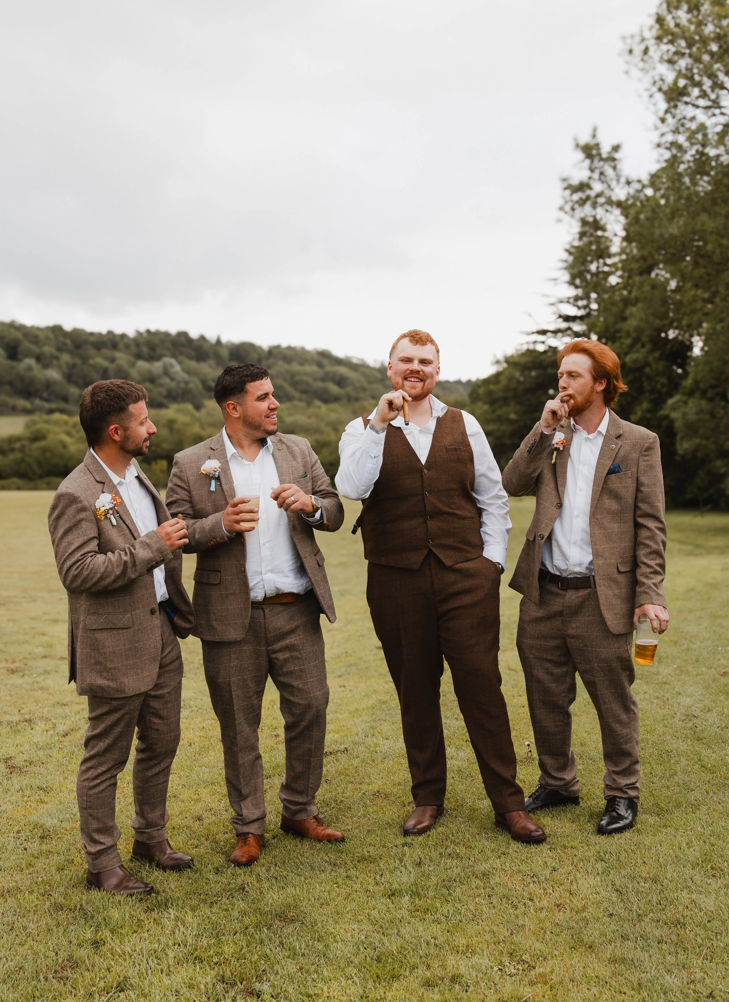 Four men dressed in suits standing on grass outdoors, drinking beer, smiling, and talking, with trees and hills in the background.