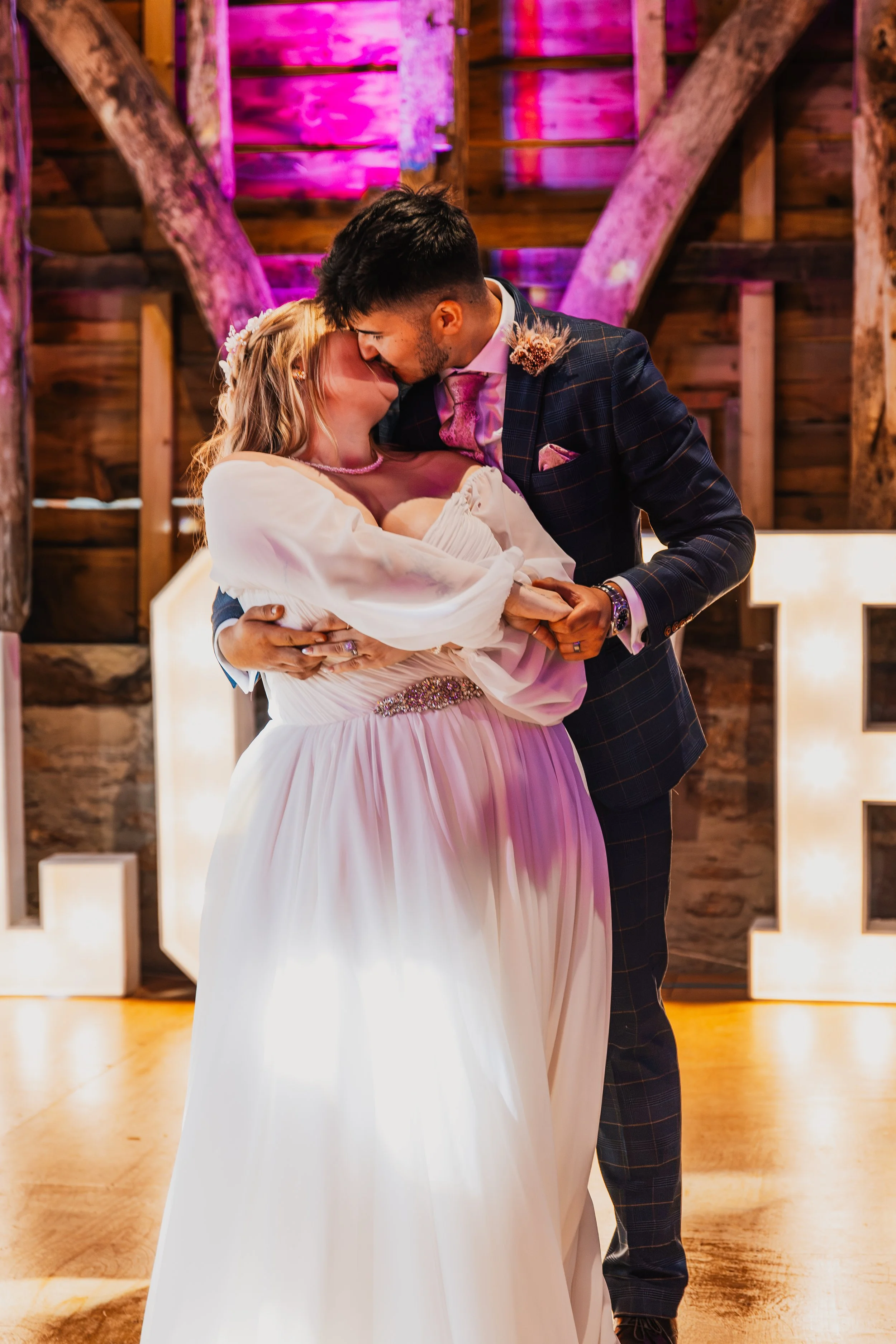 A bride and groom sharing a kiss during their wedding reception in a rustic barn decorated with pink lighting.