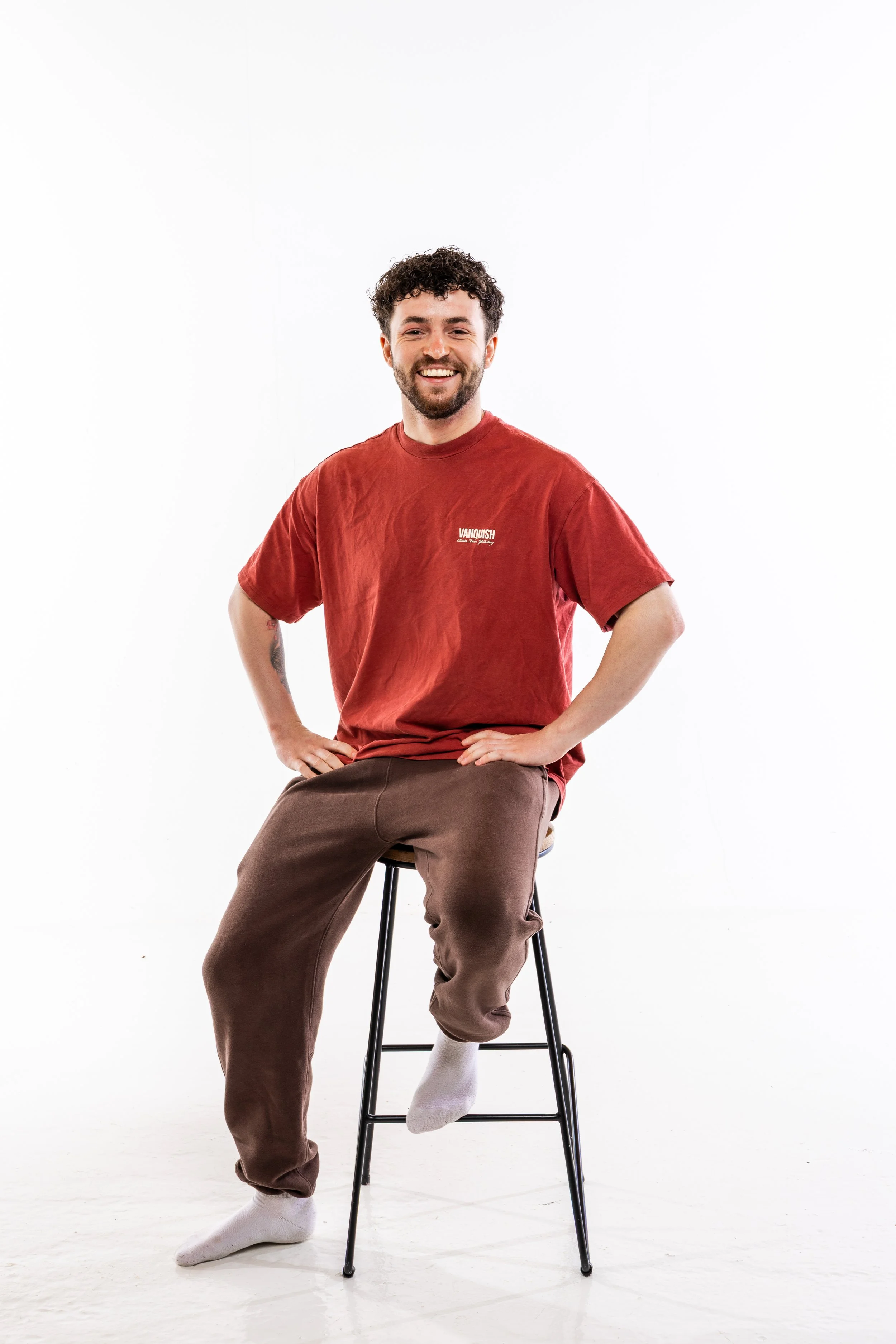 A smiling man with curly hair and a beard sitting on a stool, wearing a red T-shirt with the word 'Vancouver' on it, brown pants, and white socks, against a plain white background.