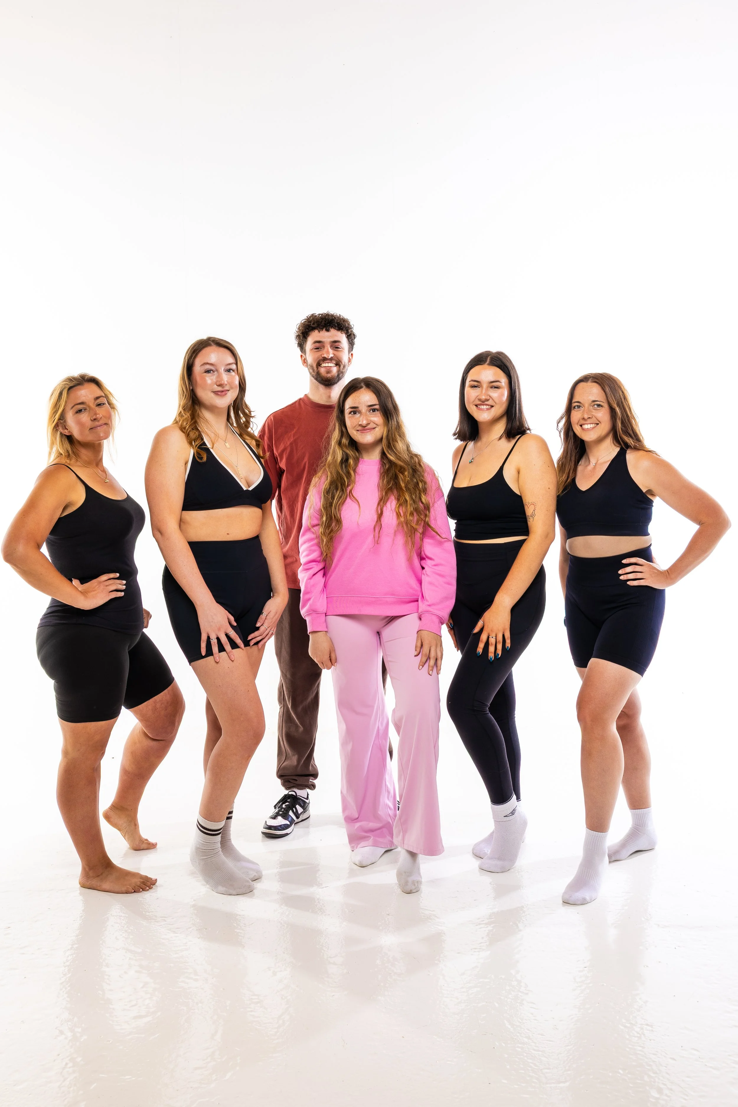 A group of six diverse young adults, five women and one man, standing together in a studio with a white background, smiling and posing for a photo. They are dressed in sportswear and casual clothing, with some wearing socks and standing barefoot.