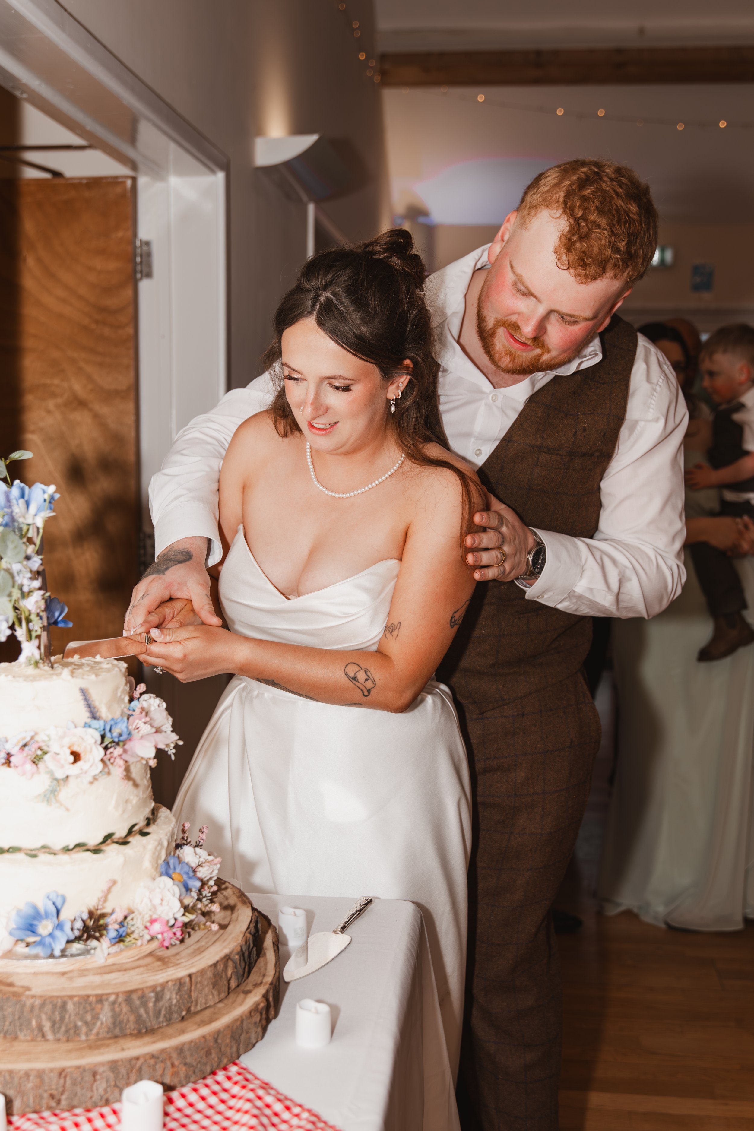 A woman in a white dress and pearl necklace is cutting a wedding cake. She is standing in front of a large, tiered cake decorated with flowers, placed on a wooden slab. A man in a white shirt and brown vest is standing behind her, placing his hand on