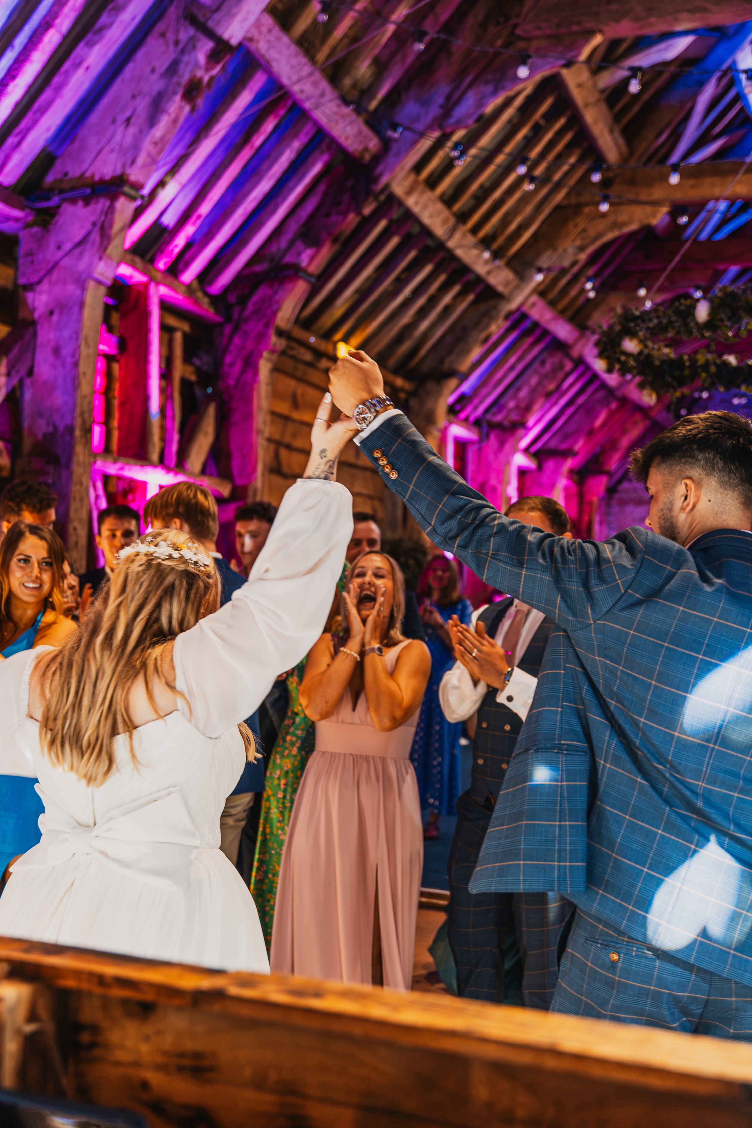 Group of people dancing at a wedding reception inside a rustic barn decorated with string lights and purple uplighting. The bride and groom are visible, with the groom raising his hand. Guests are smiling and enjoying the celebration.