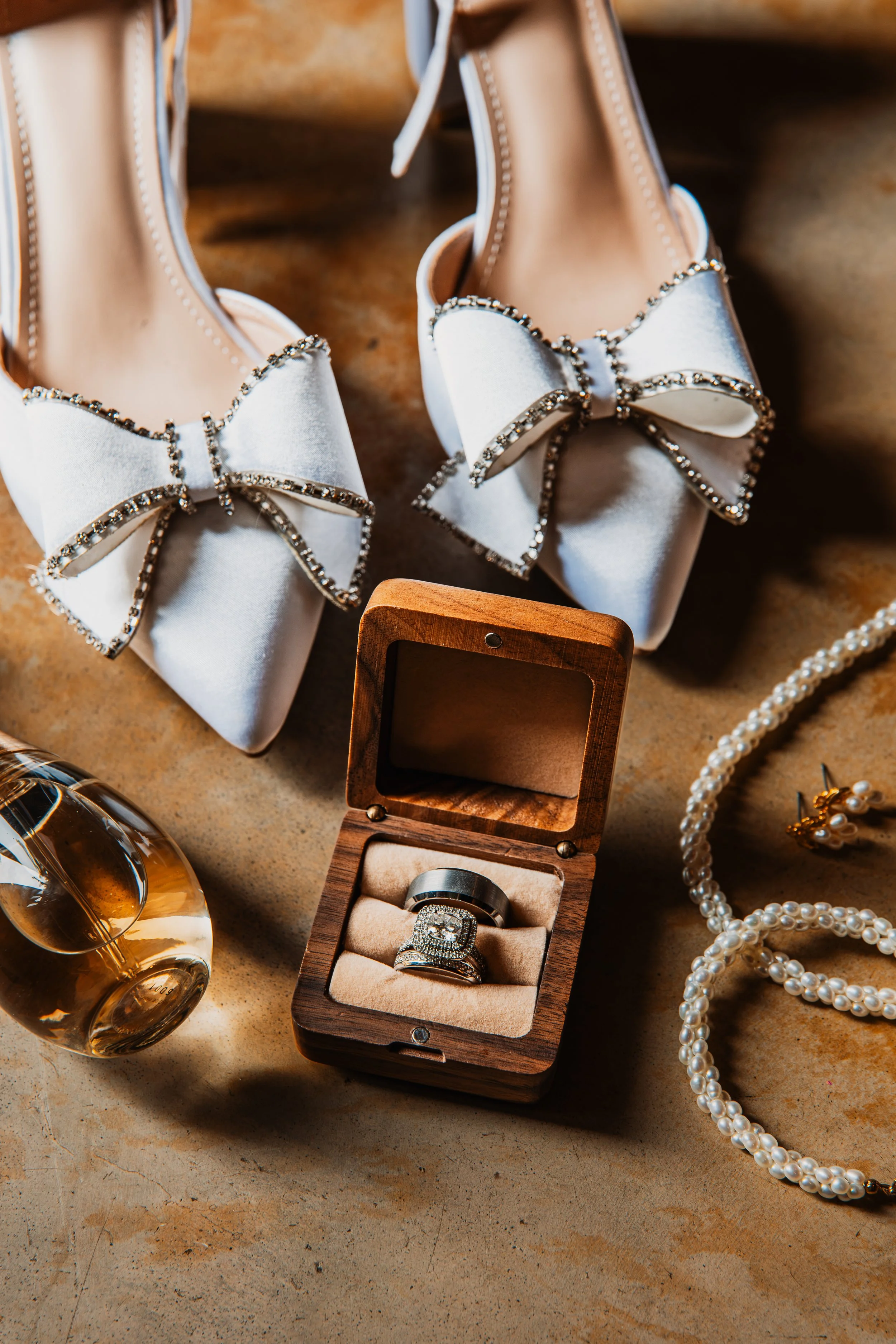 Close-up of white satin shoes with large bows encrusted with rhinestones, a wooden box with diamond rings, pearl necklace, and a glass of amber-colored drink on a textured surface.