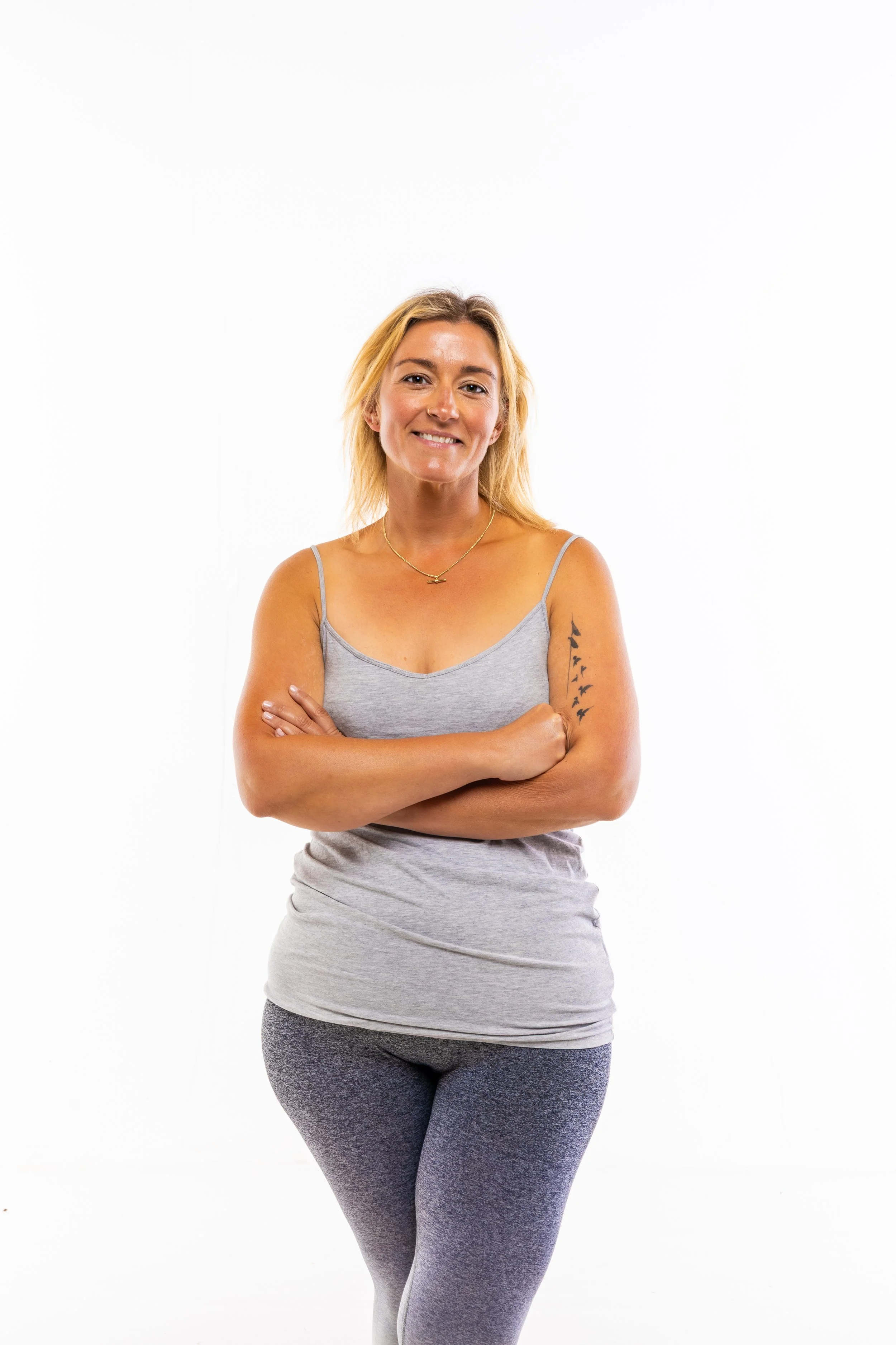 A woman with blonde hair and a tattoo on her left arm is smiling with her arms crossed, wearing a light gray tank top and dark gray leggings, standing against a white background.