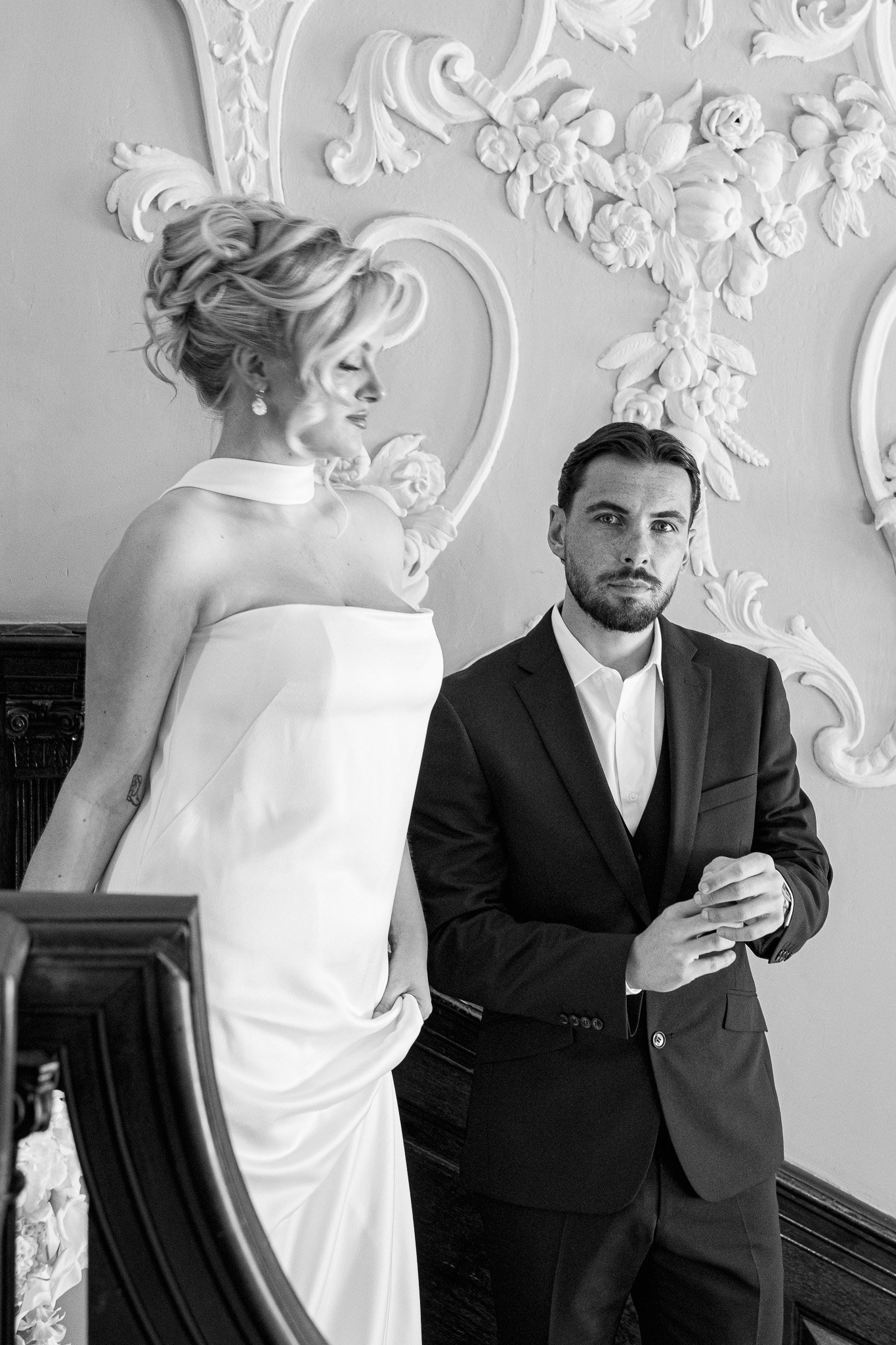 A woman in a strapless wedding gown with a choker necklace and styled blonde hair, and a man in a suit with a white shirt, sit in a decorated room with floral and ornate wall details.