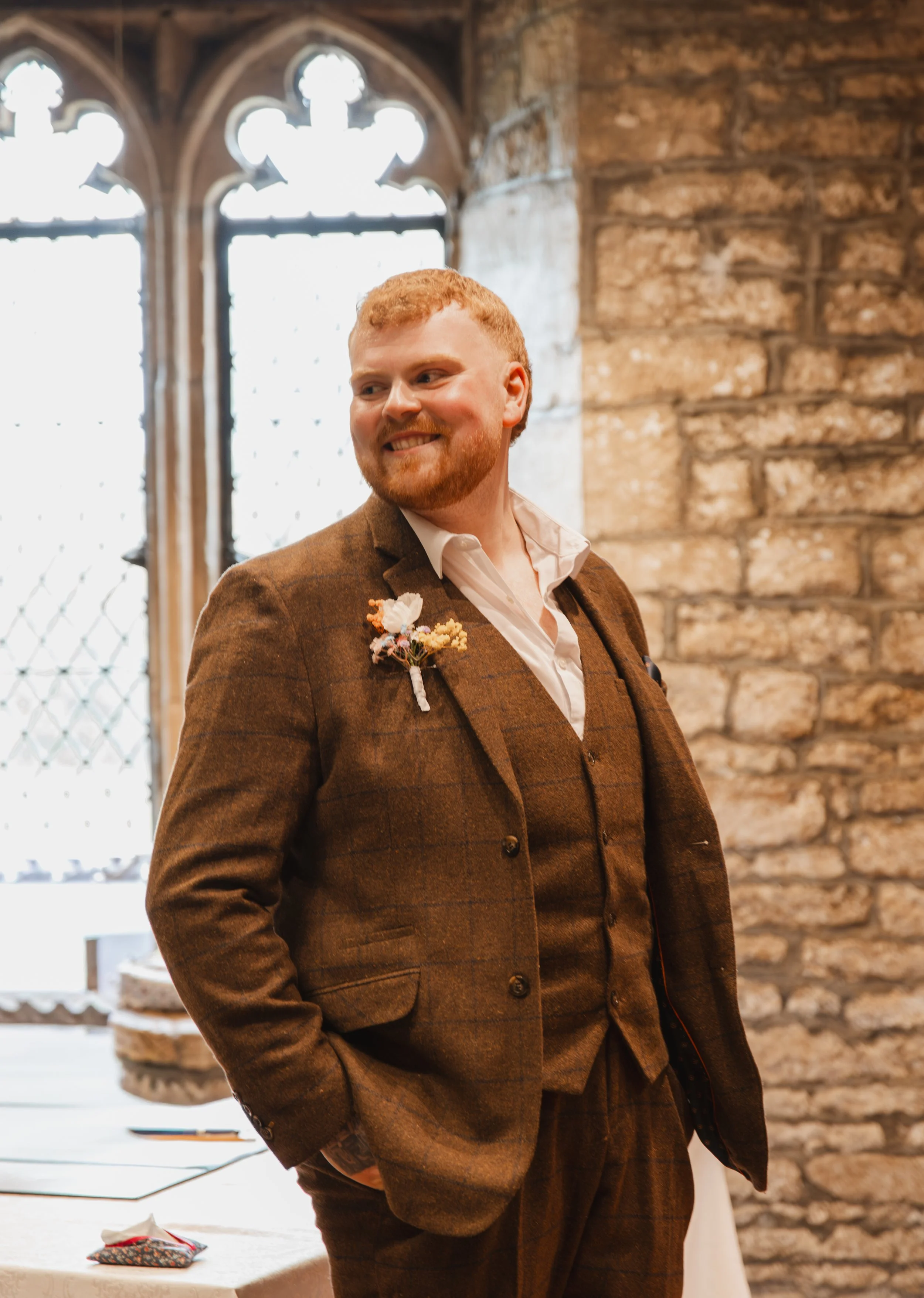 A man with red hair and beard, wearing a brown plaid suit and white shirt, smiling in an indoor historic stone building with large arched windows.
