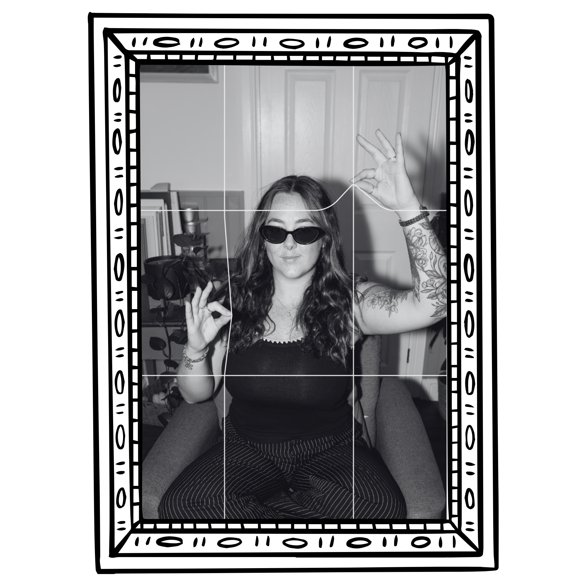 A woman with long wavy hair wearing sunglasses, black sleeveless top, and striped pants, seated indoors on a sofa, making an OK gesture with her right hand and holding a small object in her left hand, with a bookshelf and door visible in the background.