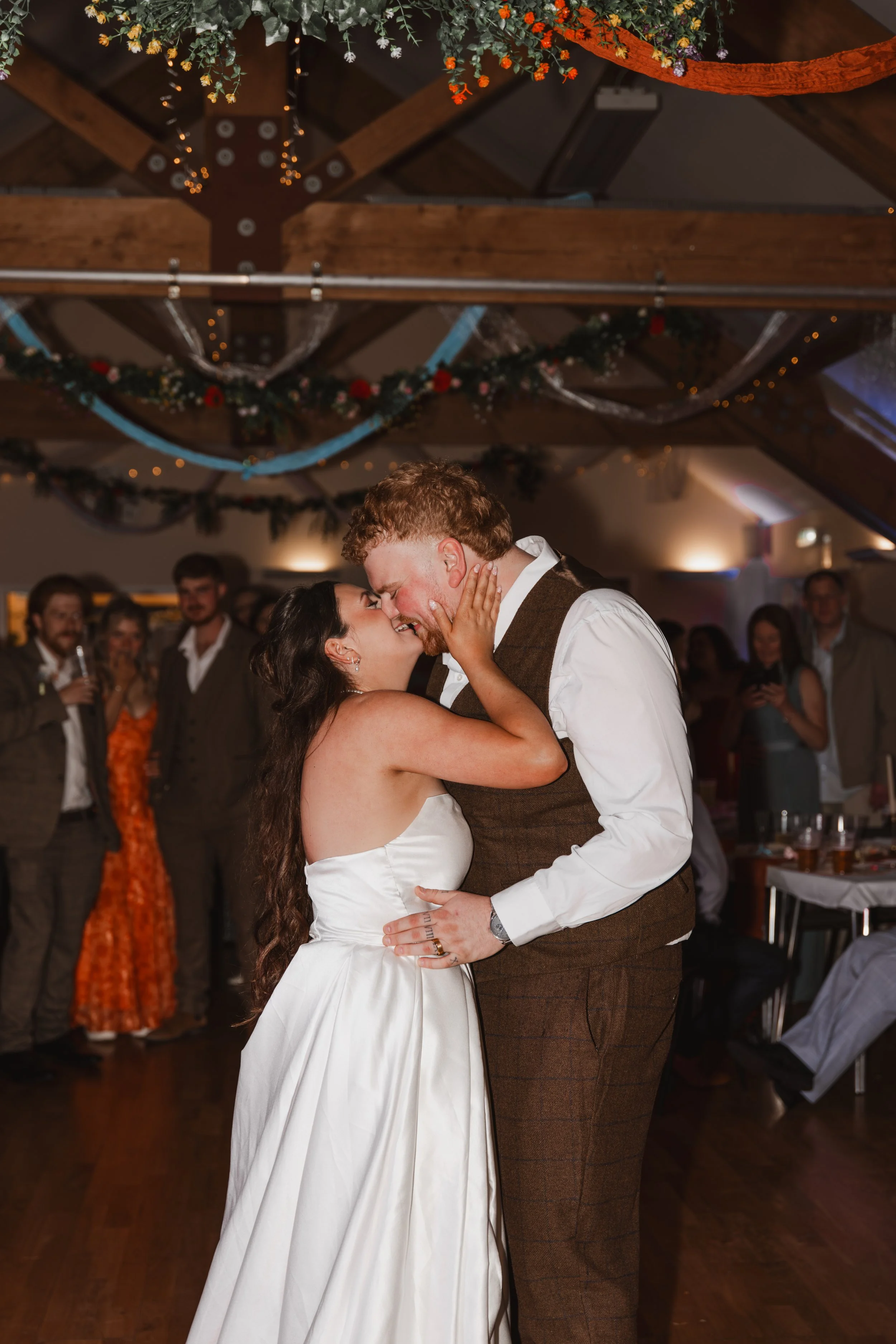 A bride and groom share a dance at their wedding reception, surrounded by guests in a decorated venue with hanging floral arrangements and string lights.