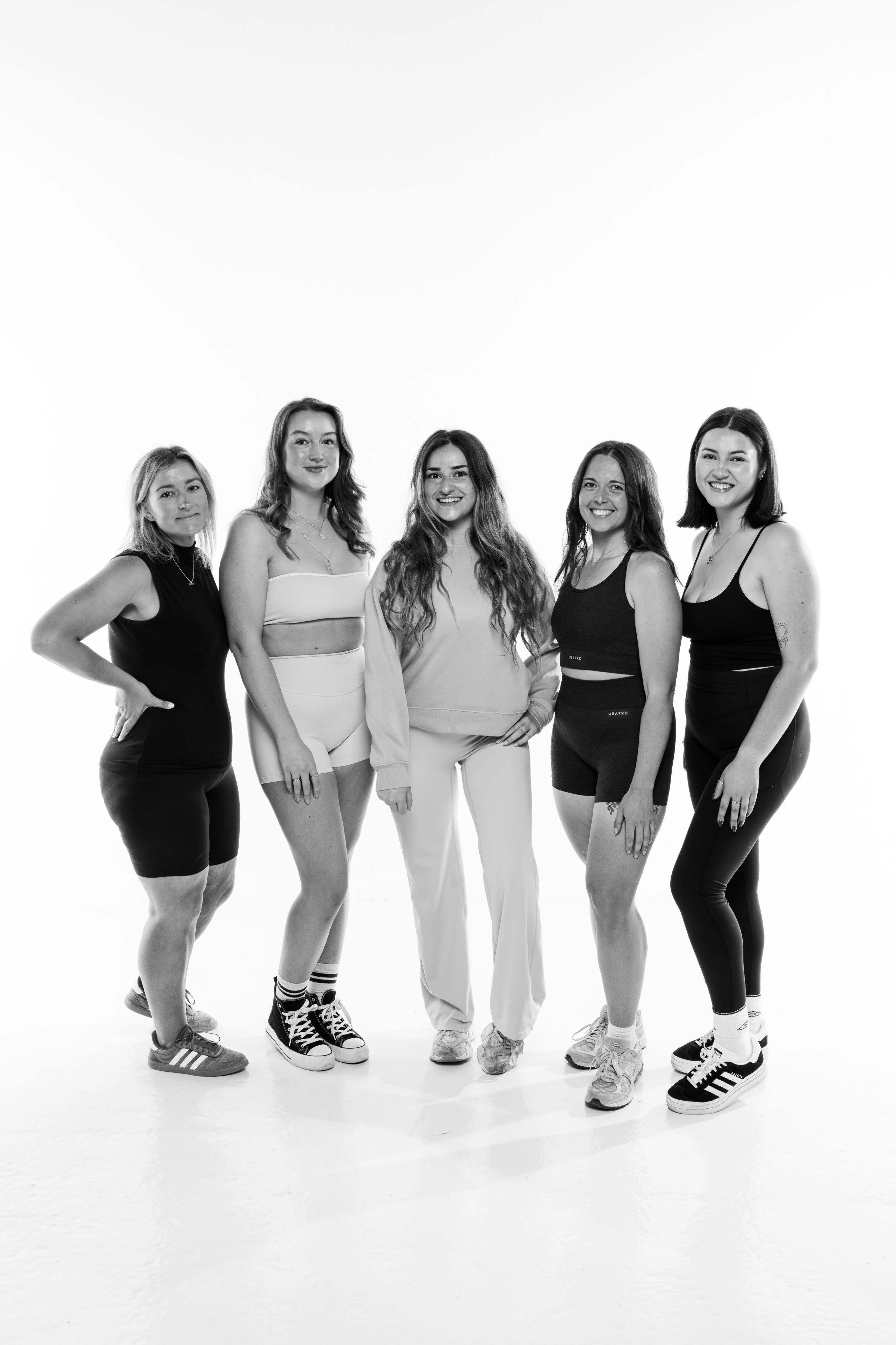A group of five women standing together in casual athletic clothing against a plain white background.