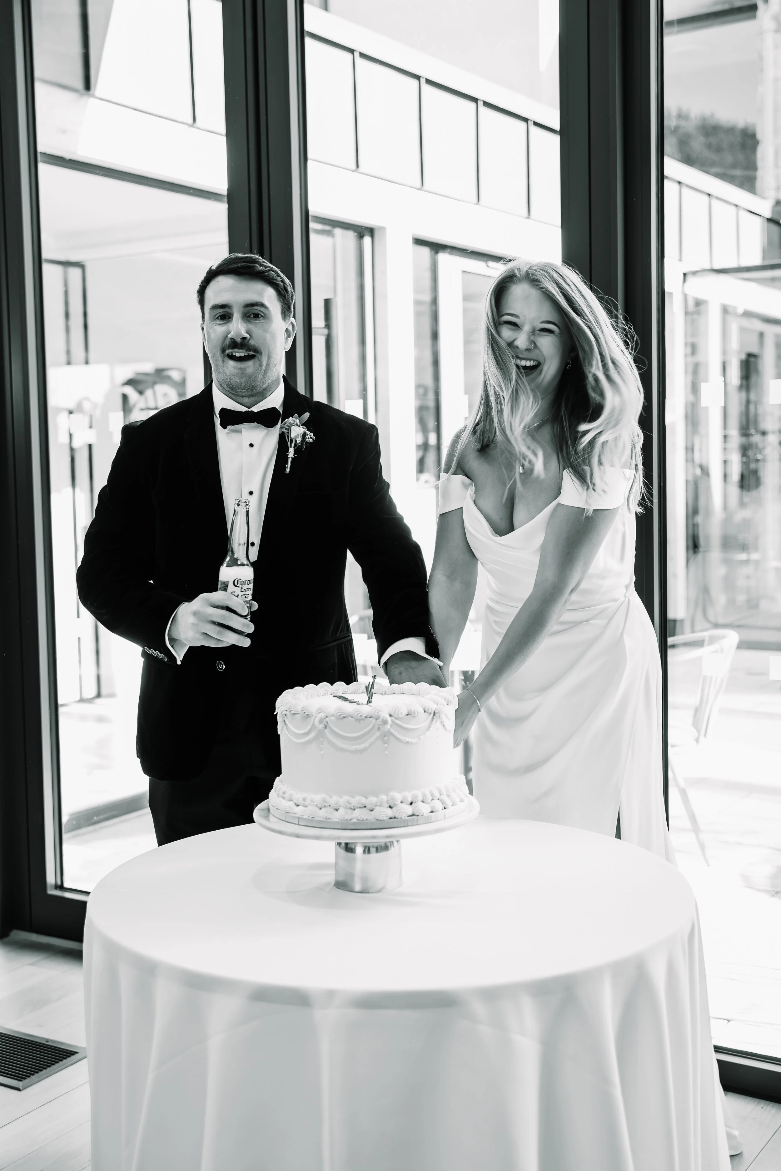 A black-and-white photo of a bride and groom cutting a wedding cake at a reception, with the groom holding a beer bottle, in a modern indoor venue with glass windows.