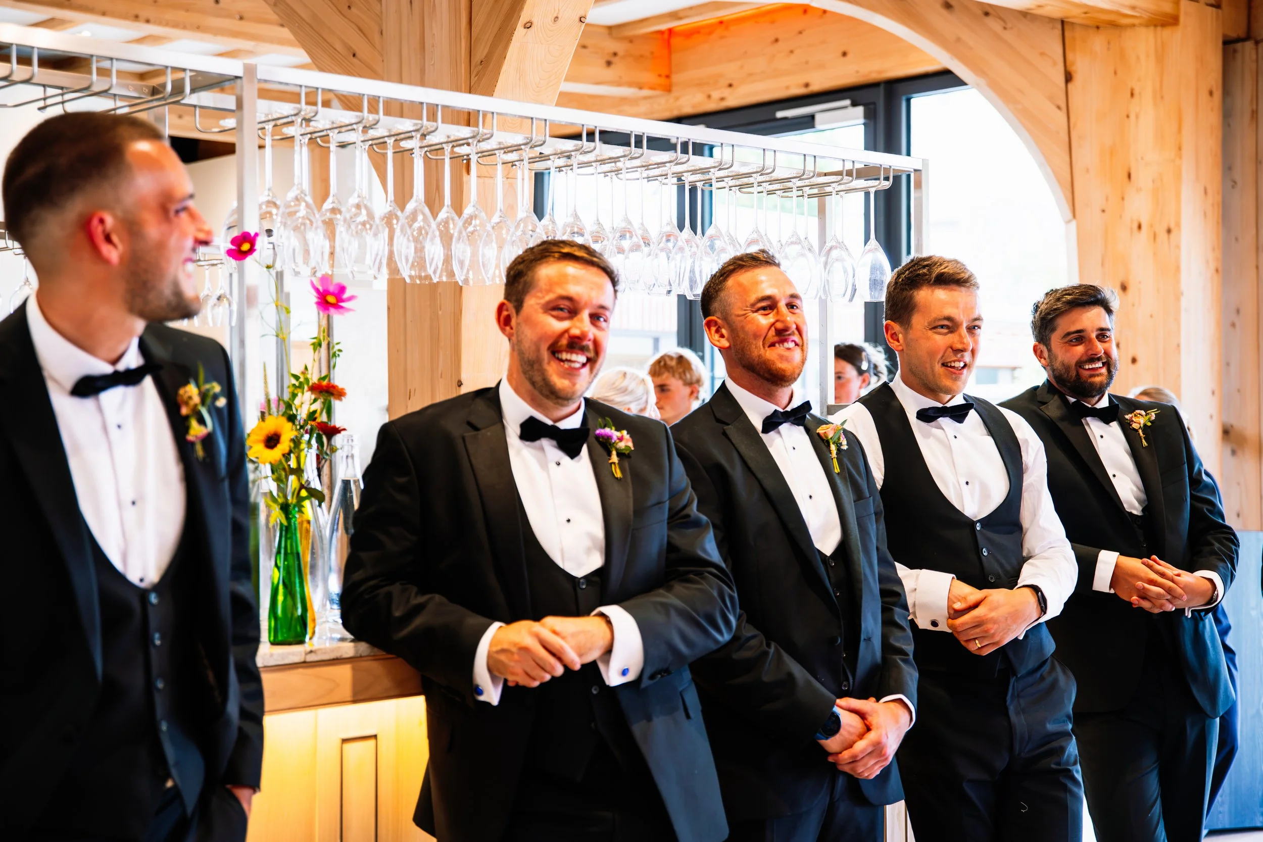Groom and groomsmen dressed in black tuxedos with bow ties, standing in a row at a wedding reception inside a wooden building, with wine glasses hanging above and colorful flowers on the table.