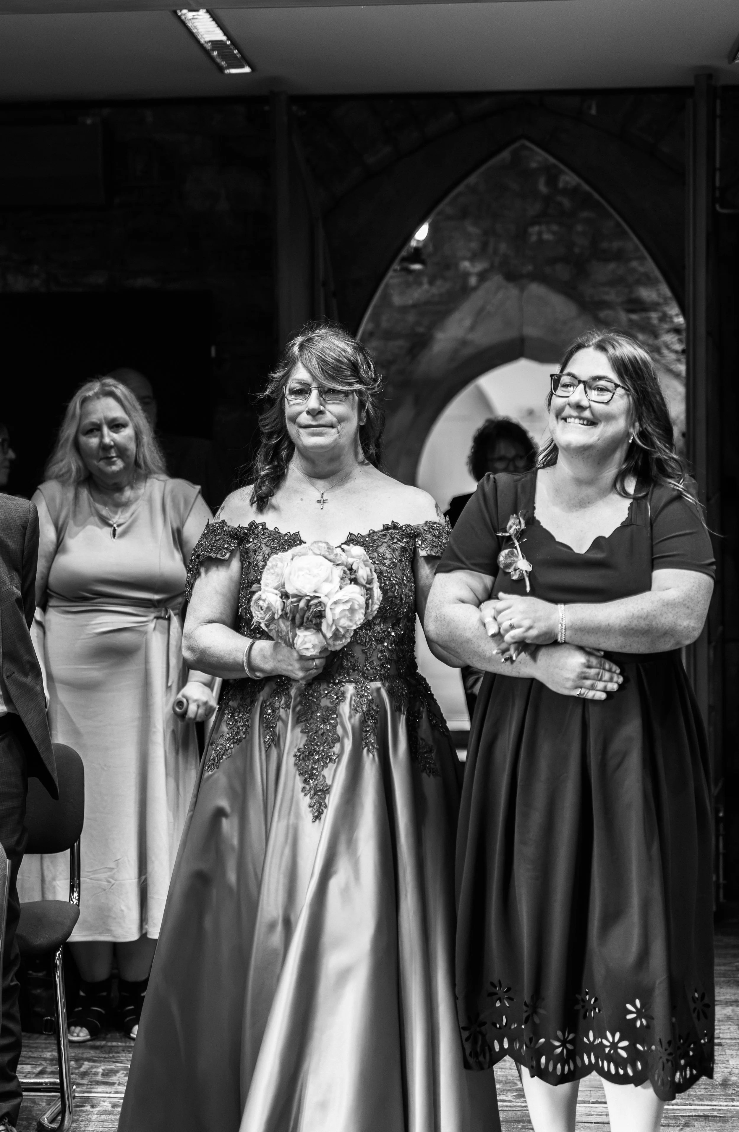 A group of women dressed in formal attire attending a celebratory event. The woman in the center is holding a bouquet of flowers and wearing glasses, an off-shoulder dress with intricate embroidery. Other women are standing nearby, some smiling and o
