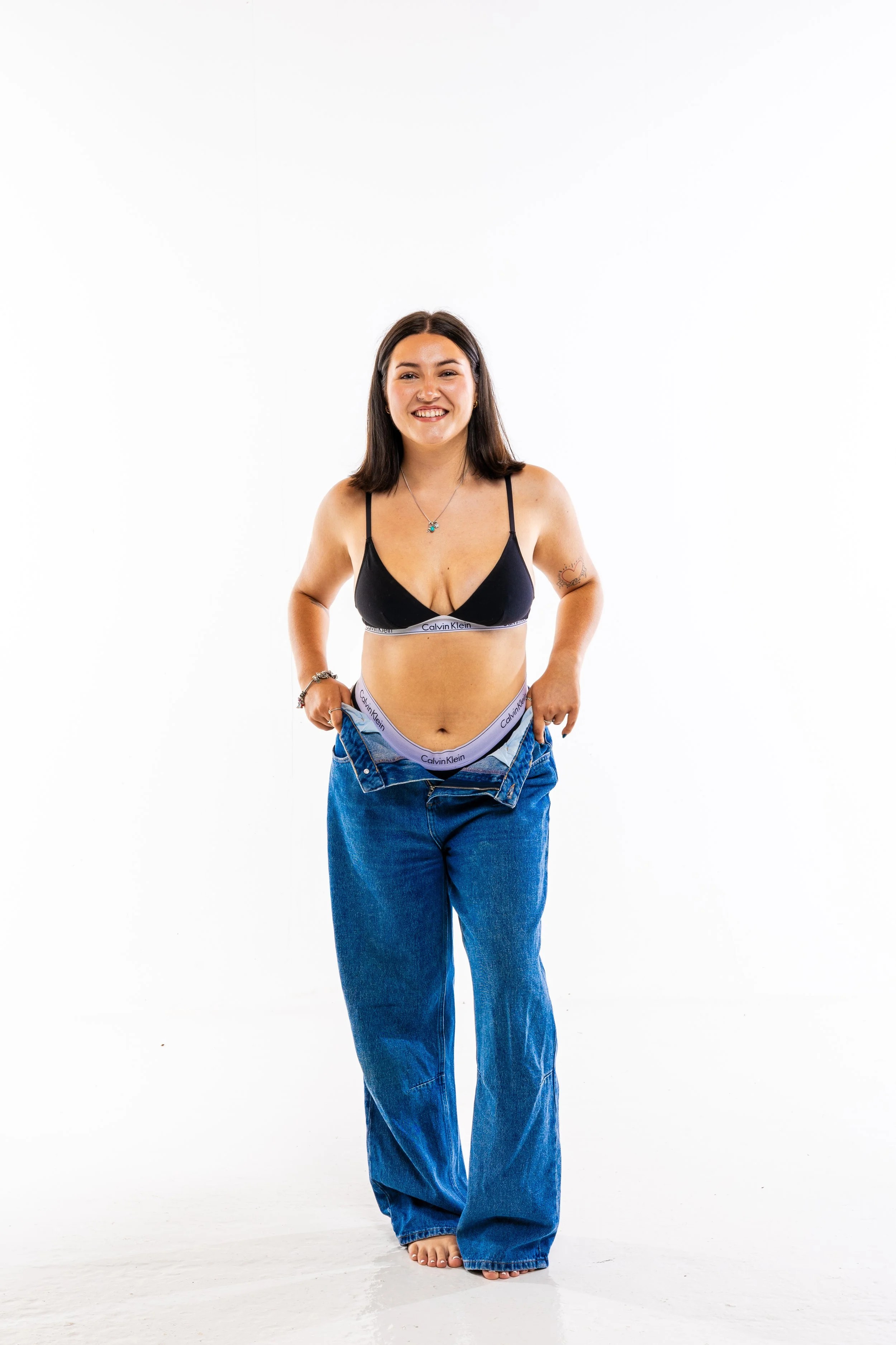 Young woman standing barefoot against a white background, wearing loose blue jeans, Calvin Klein underwear, and a black bralette, smiling at the camera.