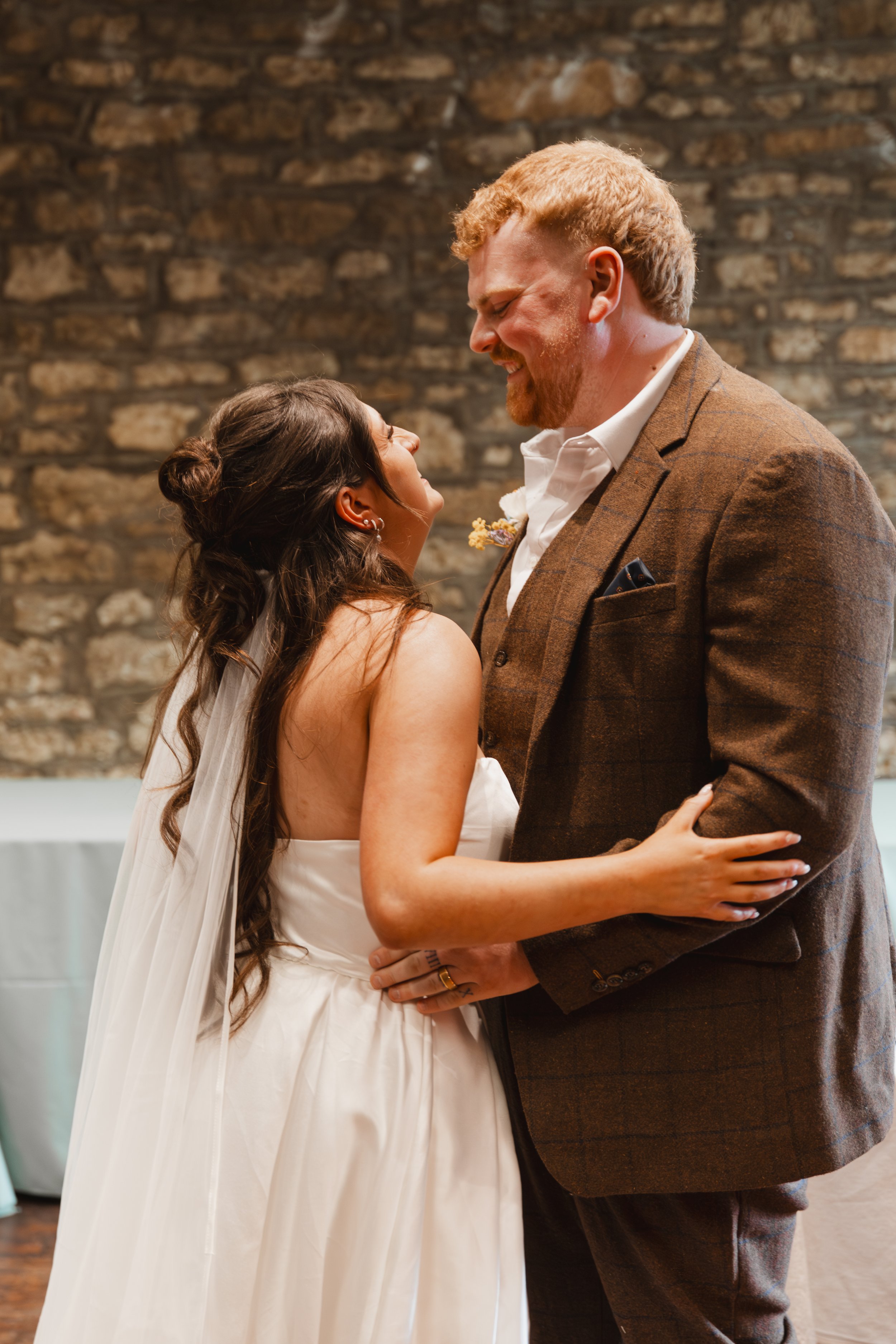 A bride and groom are dancing together at their wedding, gazing into each other's eyes and smiling, with a stone wall in the background.