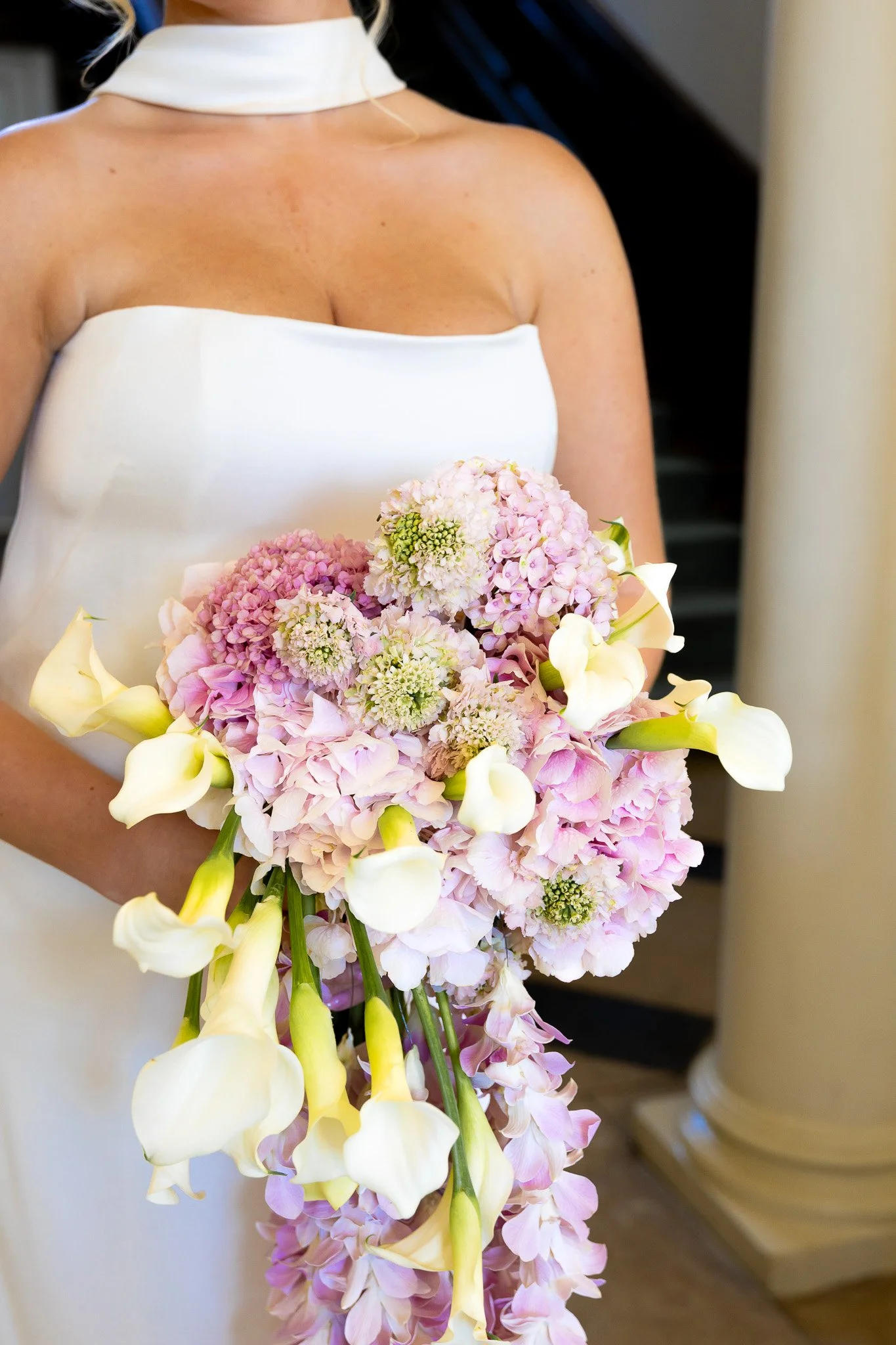 A woman in a white strapless dress holding a large bouquet of pink and white hydrangeas and calla lilies.