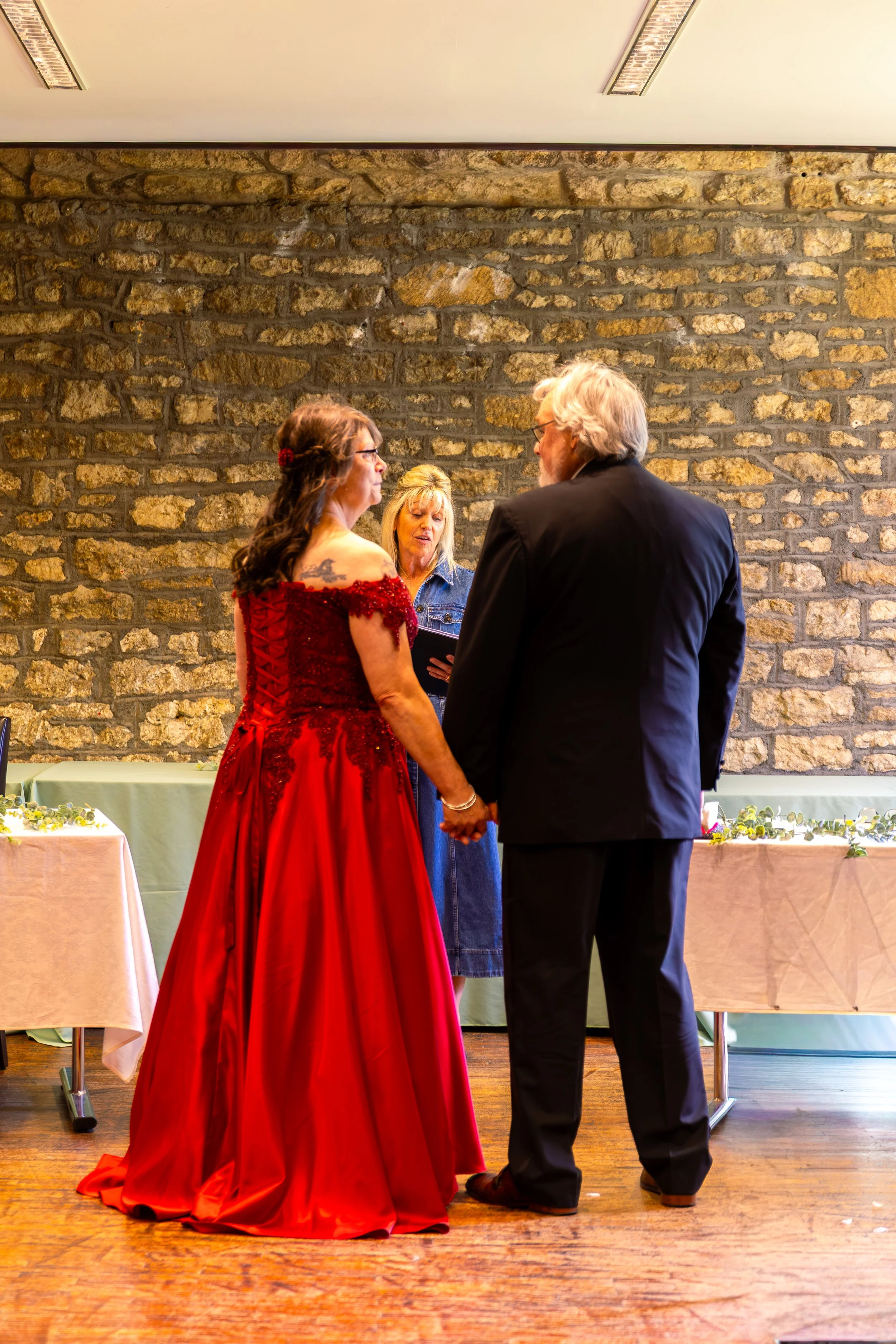 A couple is getting married, holding hands while standing before an officiant. The woman is wearing a red, off-the-shoulder gown, and the man is in a black suit. They are in a room with a brick wall background and tables with tablecloths.