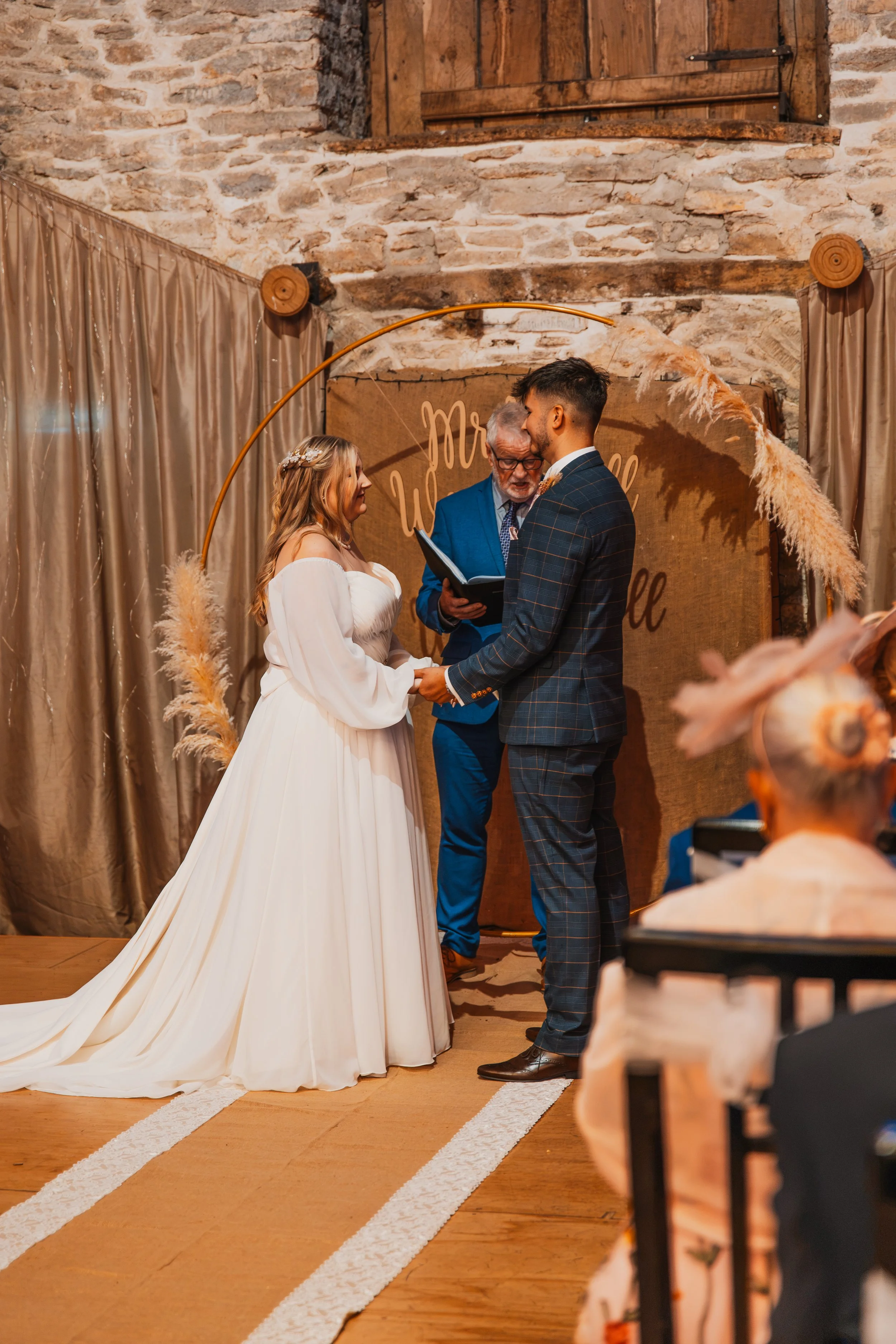 A wedding ceremony with a bride and groom holding hands, standing in front of an officiant, in a rustic indoor venue with brick walls and beige curtains. The bride wears a white dress with long sleeves, and the groom wears a plaid suit with brown sho