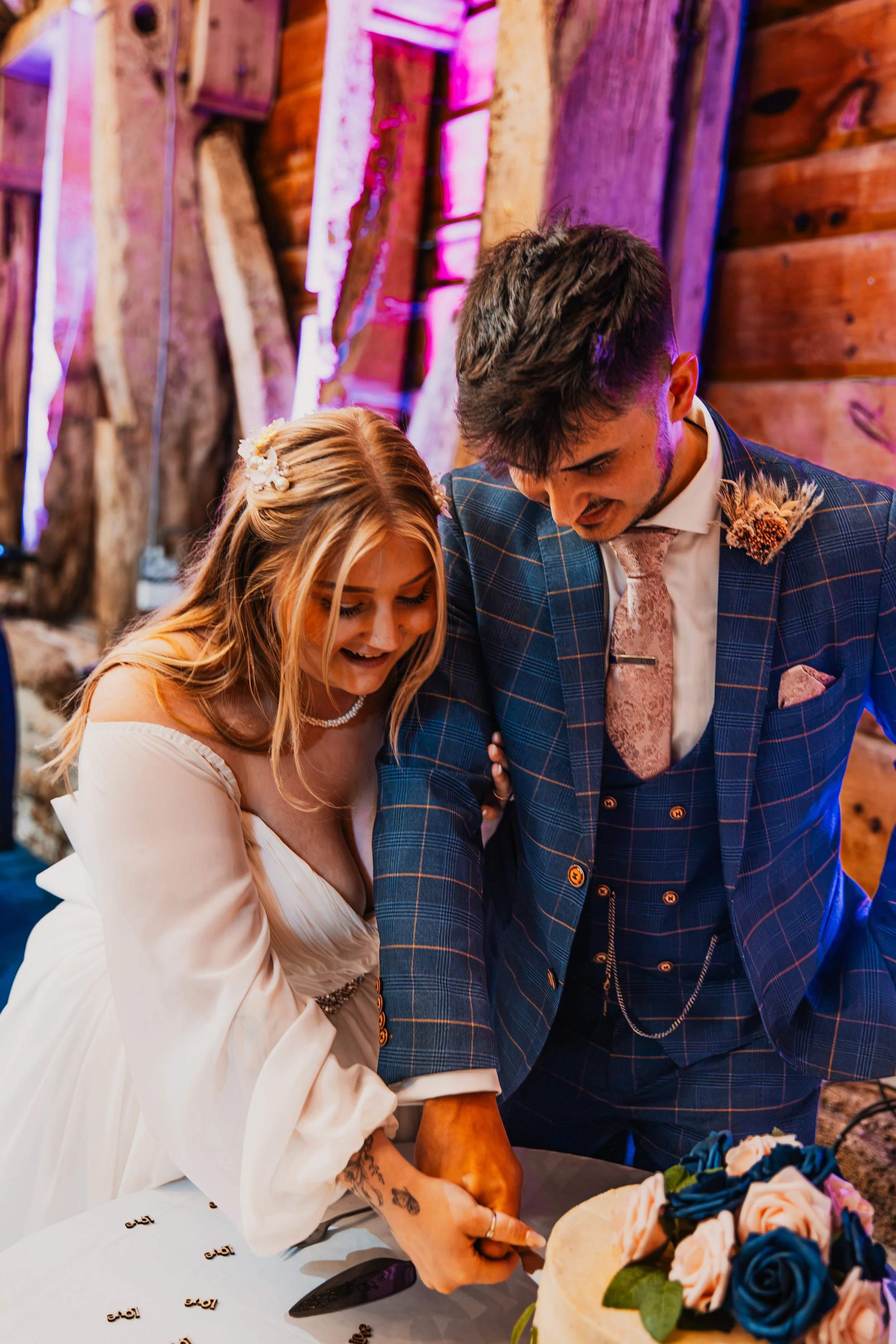 A bride and groom cutting a wedding cake together at their wedding reception. The cake is decorated with roses in shades of blue, pink, and peach, arranged with green leaves.