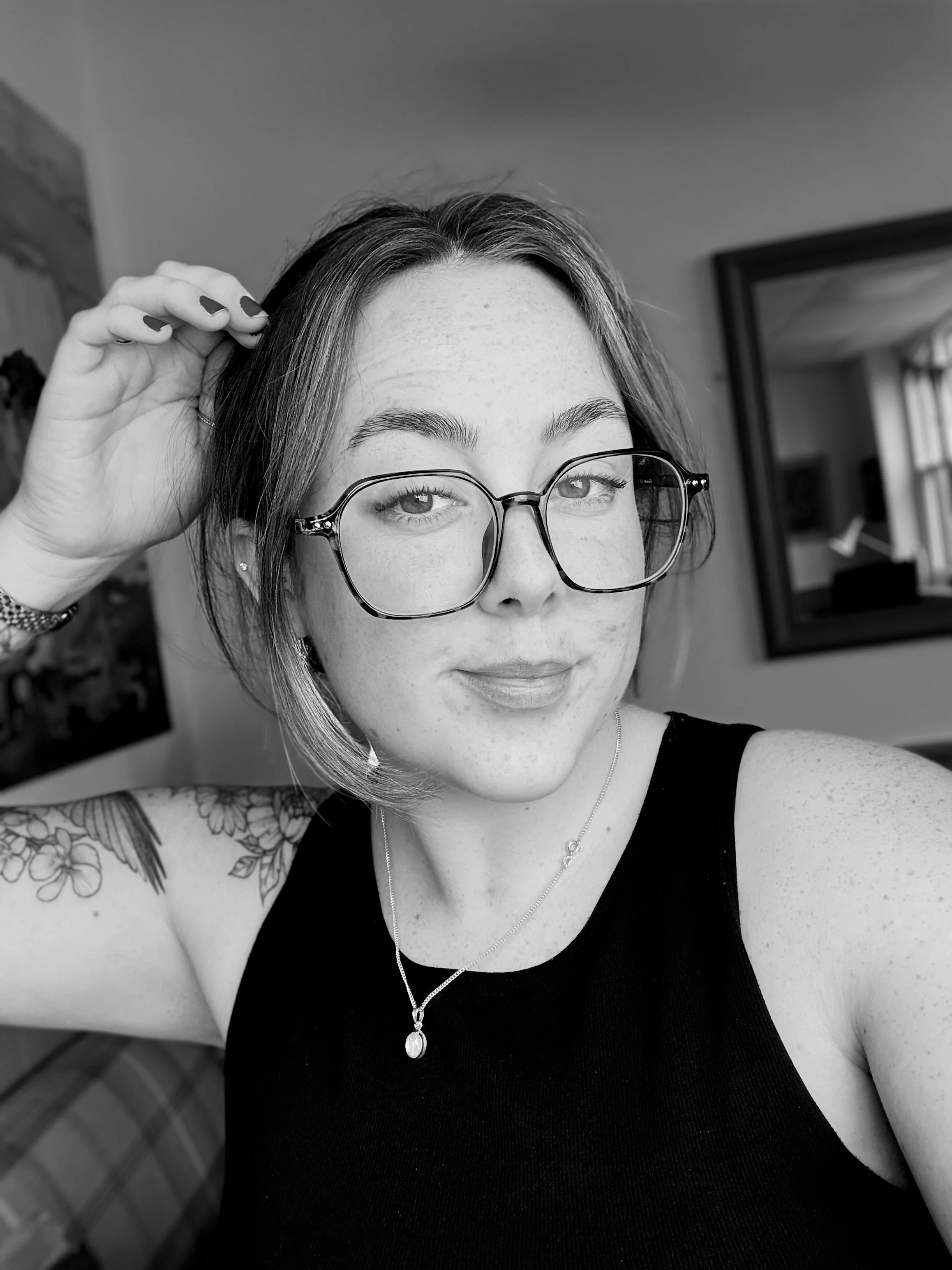 A woman with glasses taking a selfie indoors. She has short hair, freckles, a floral tattoo on her arm, and is wearing a black sleeveless top, a necklace with a pendant, and a bracelet. She is touching her hair with her right hand.