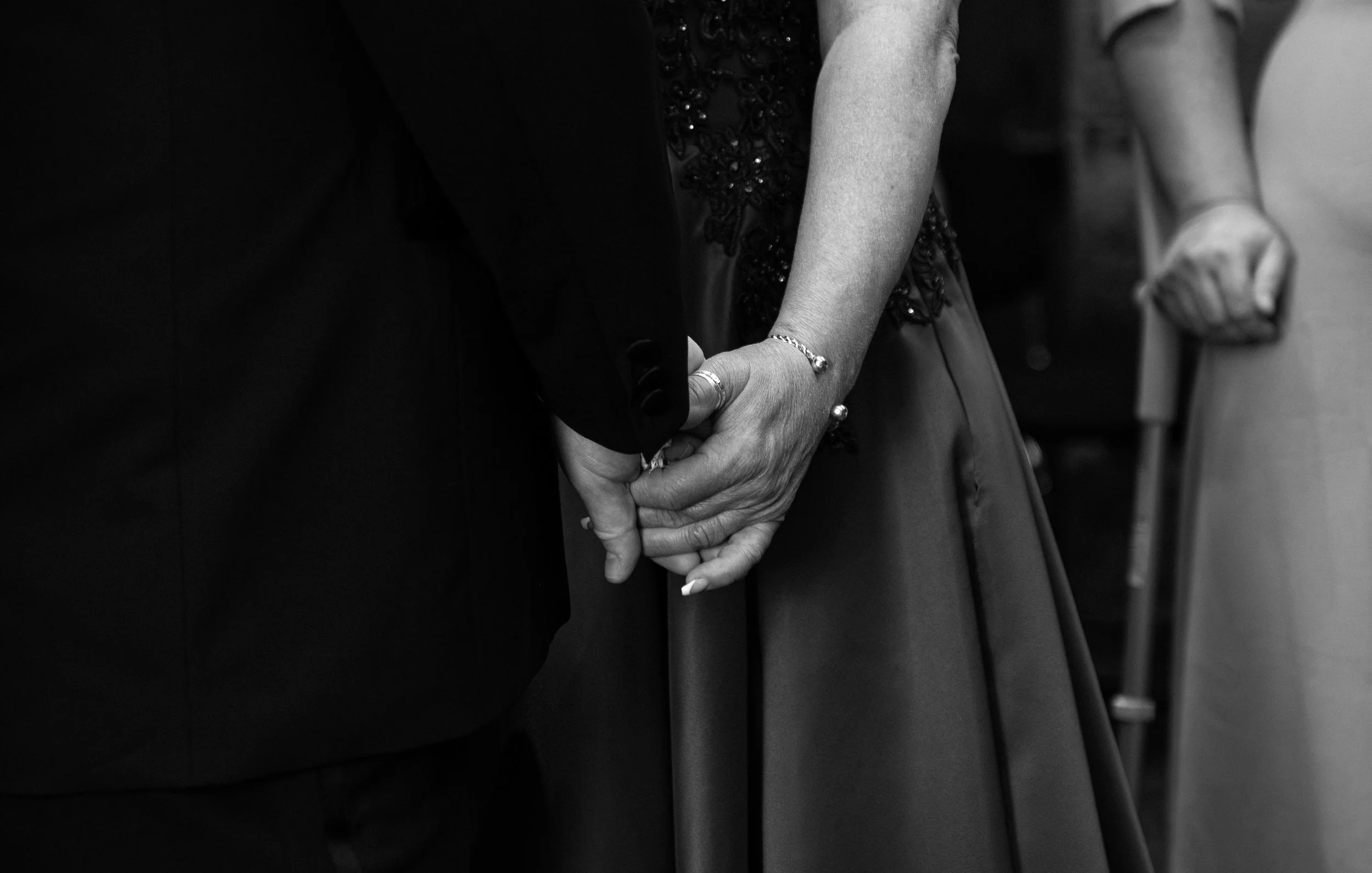 Close-up black-and-white photograph of a couple holding hands, with the woman's hand gently intertwined with the man's hand, both dressed in formal attire, indicating a romantic or special occasion.