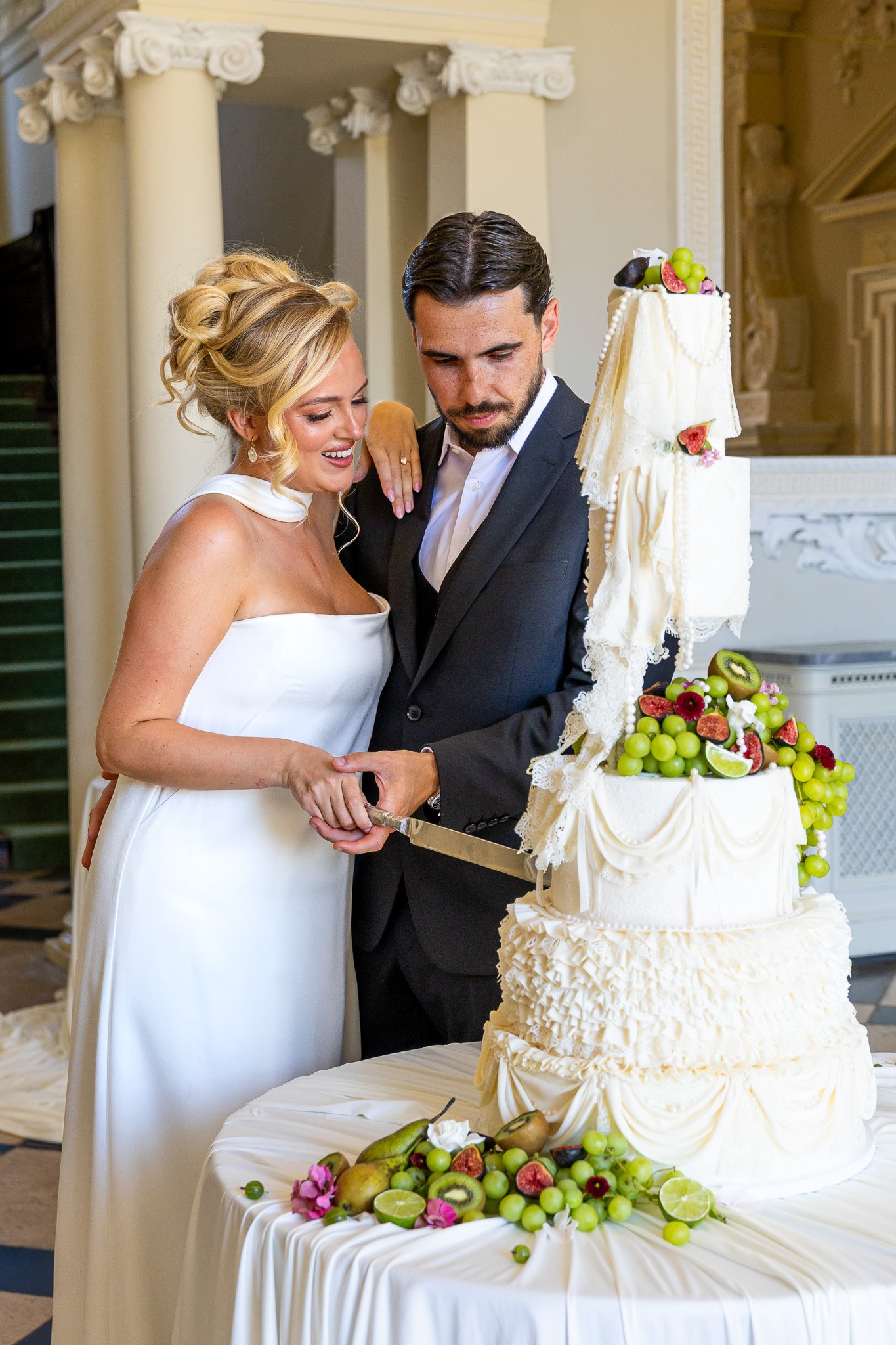 A bride and groom cutting a wedding cake decorated with white icing and fresh fruit, including strawberries, grapes, kiwis, and figs, surrounded by flowers and additional fruit on a table.