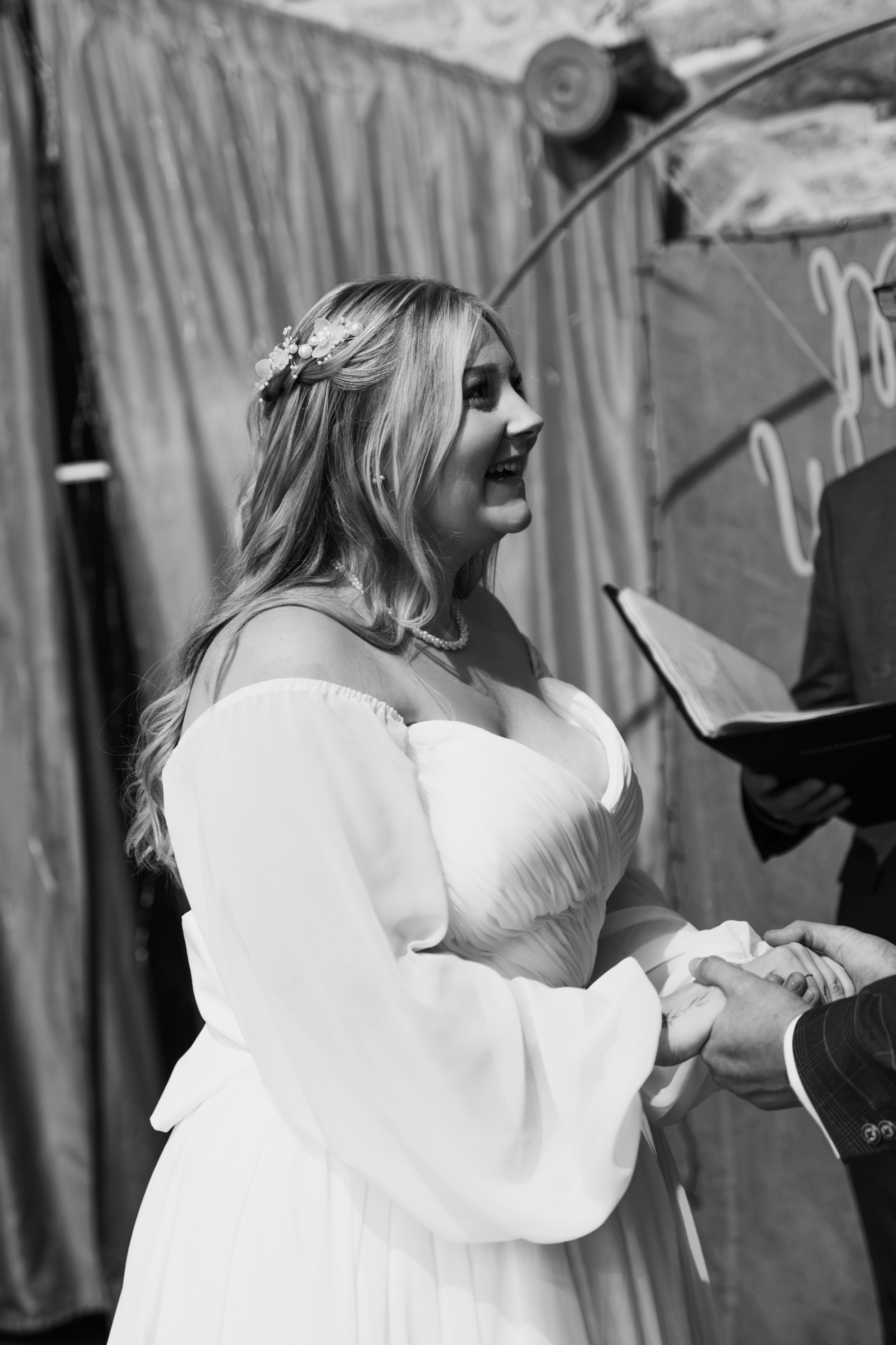 A woman with long hair in a floral headband, wearing a white dress, is smiling and holding hands with a man in a suit during a wedding ceremony.
