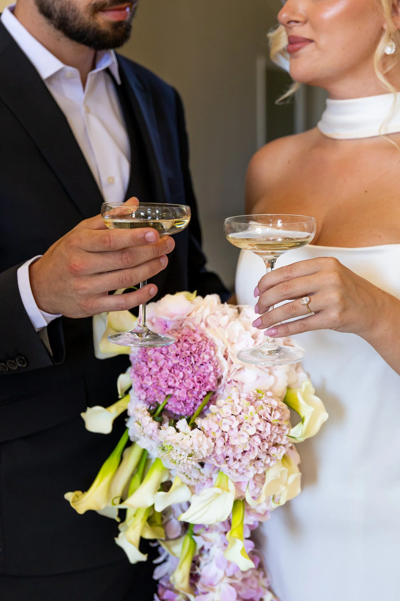 A couple at a wedding holding champagne glasses, with the bride holding a large bouquet of pink and white flowers, and the groom in a black suit and white shirt.