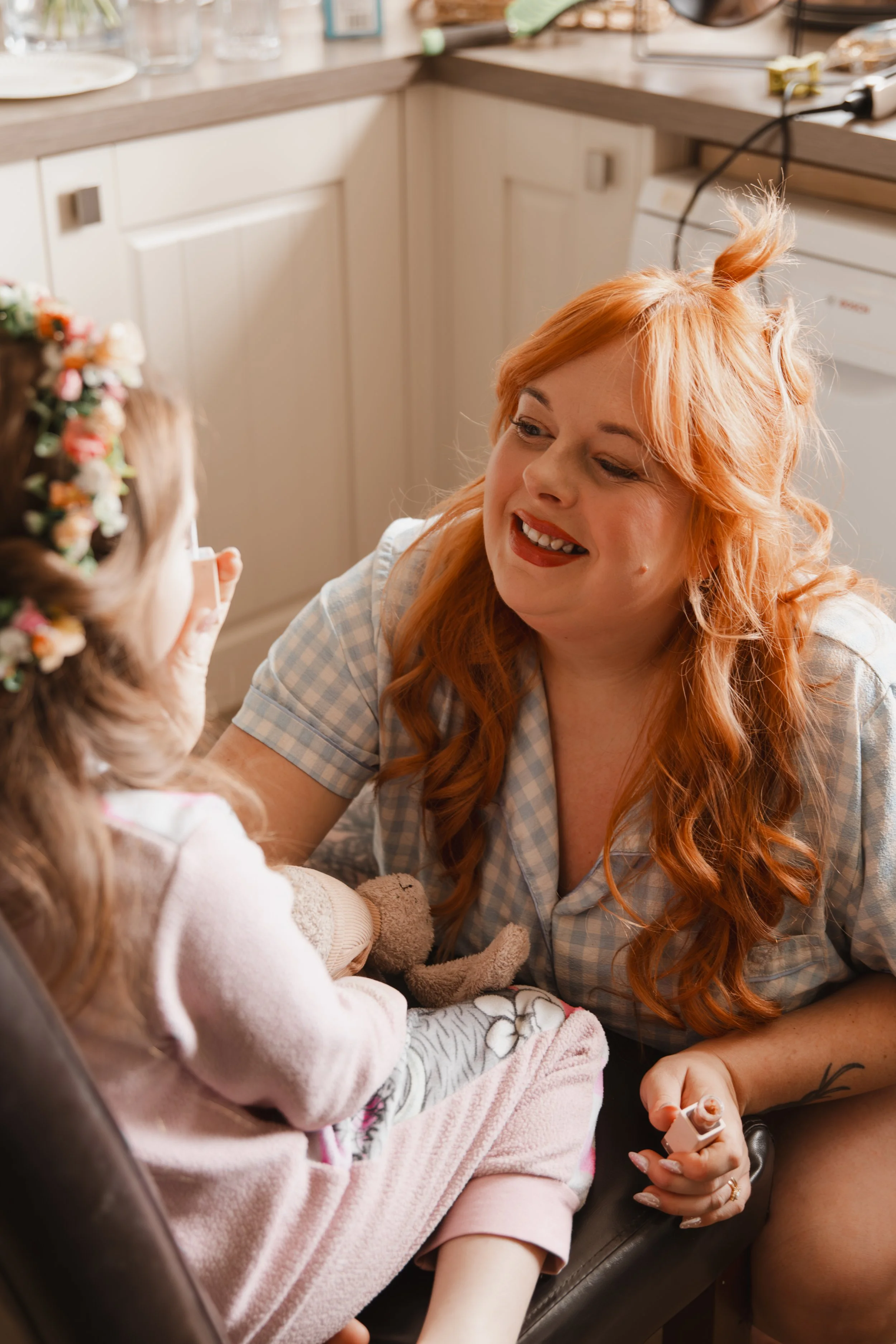 A woman with long red hair and a checkered shirt is smiling at a young girl wearing a flower crown, as they sit on the floor in a kitchen. The woman appears to be painting the girl's nails, and the girl is wearing a pink sweater and patterned legging