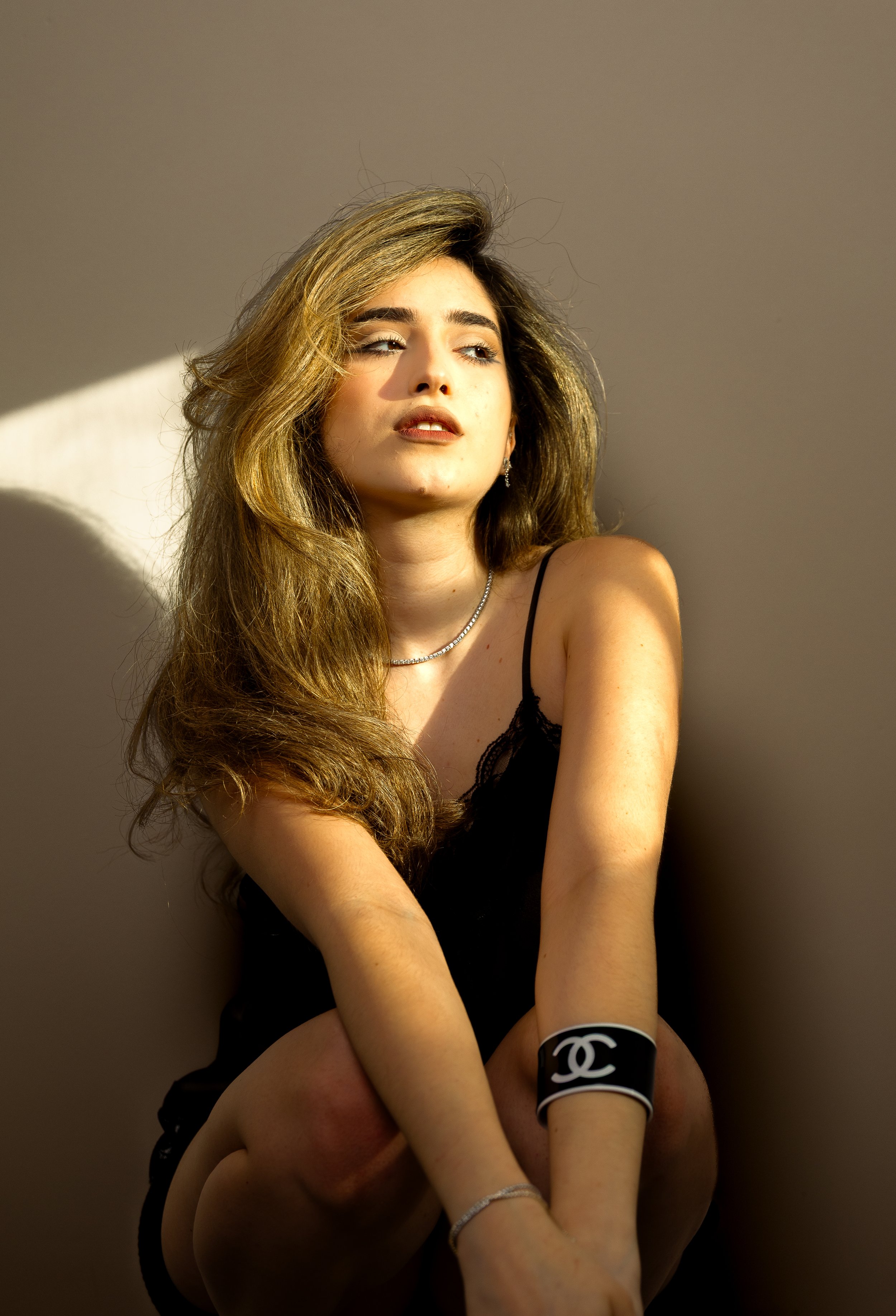 Photograph of a young woman with long, wavy light brown hair, wearing black lace clothing and jewelry, sitting against a plain wall with sunlight casting shadows.