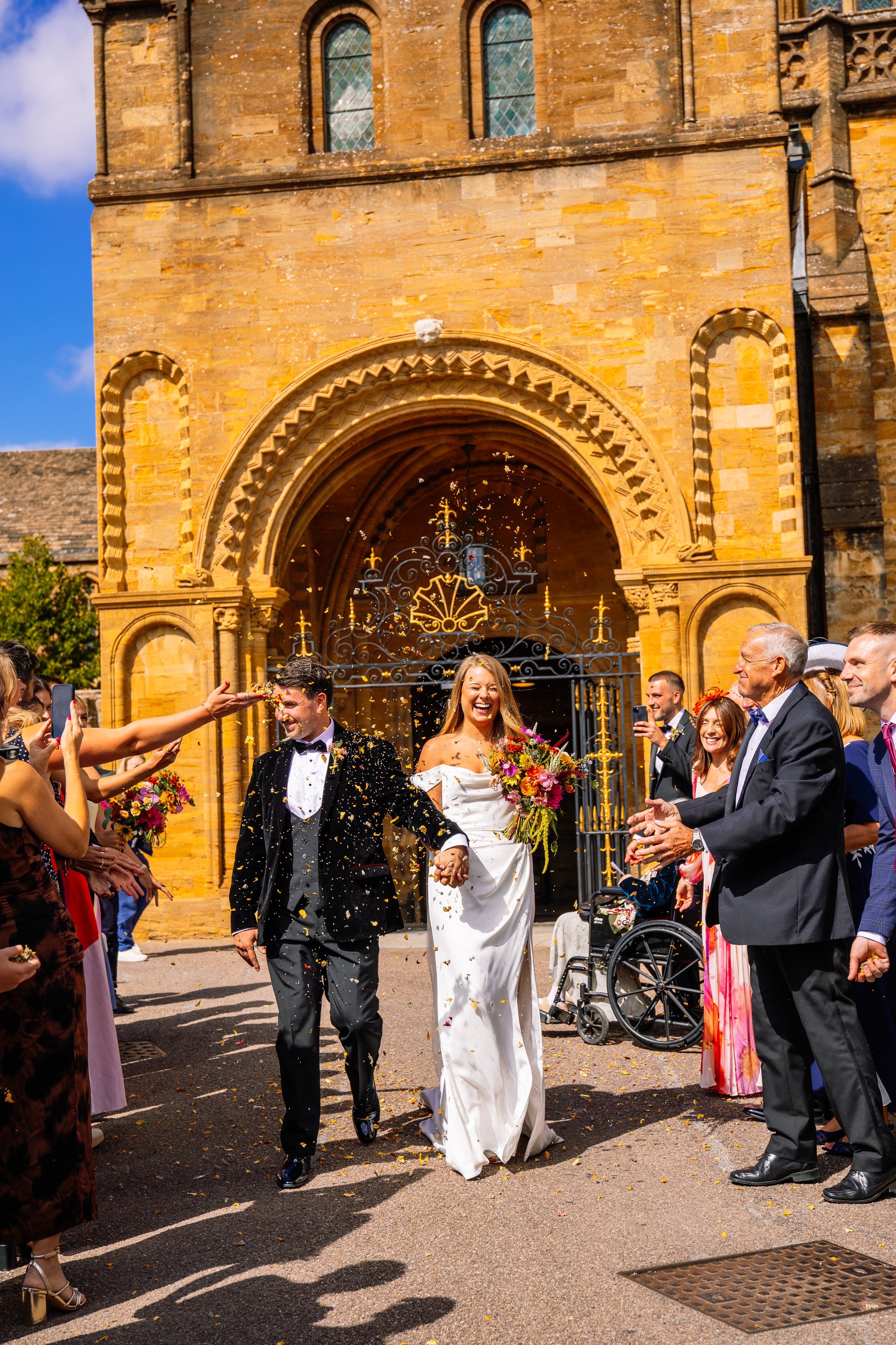 Couple in wedding attire holding hands and smiling, surrounded by guests throwing flower petals outside a church with gothic architecture in the background.