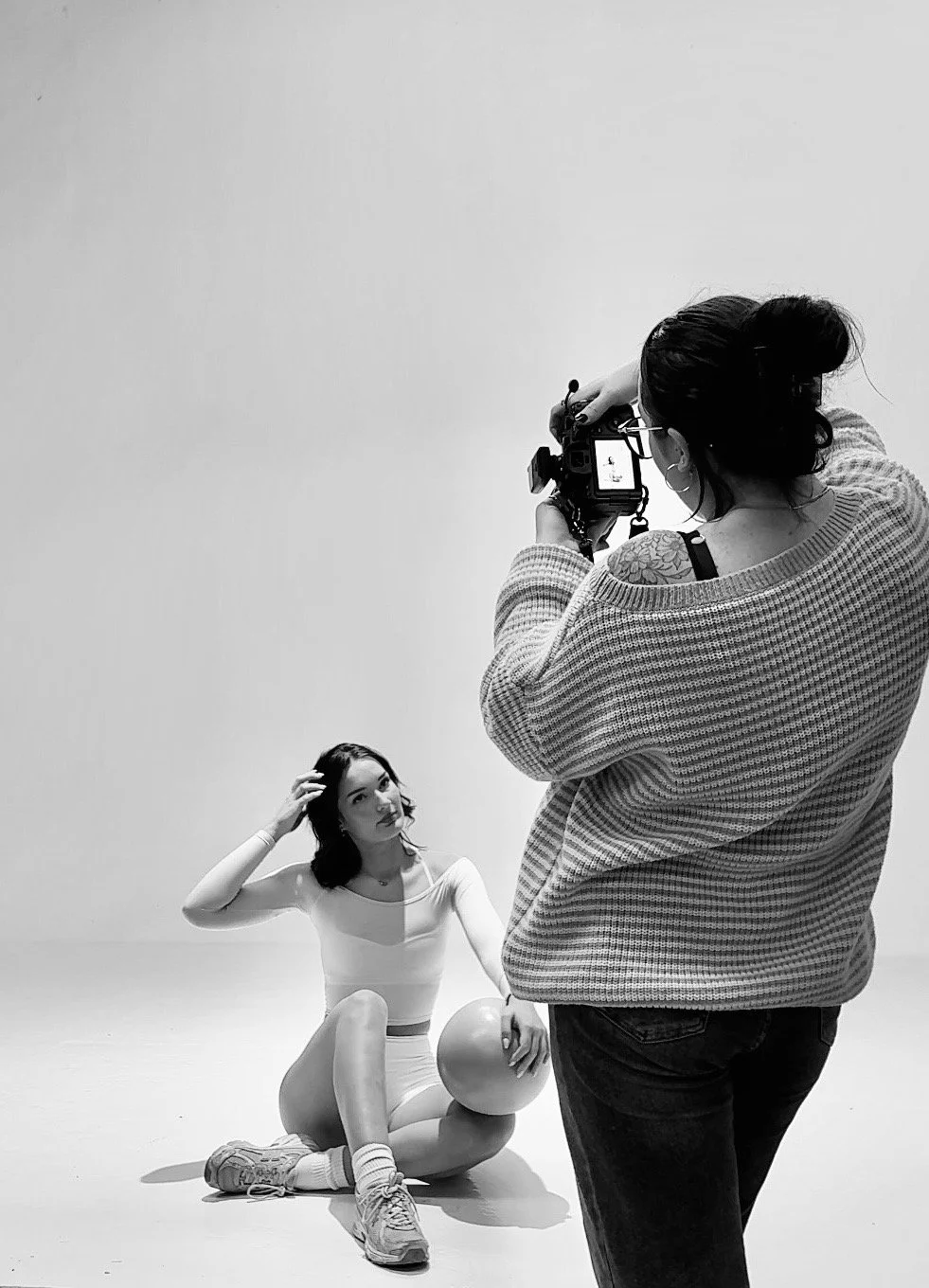 A woman with dark hair and tattoos on her shoulder takes a photo of a seated woman holding a balloon, against a plain backdrop.