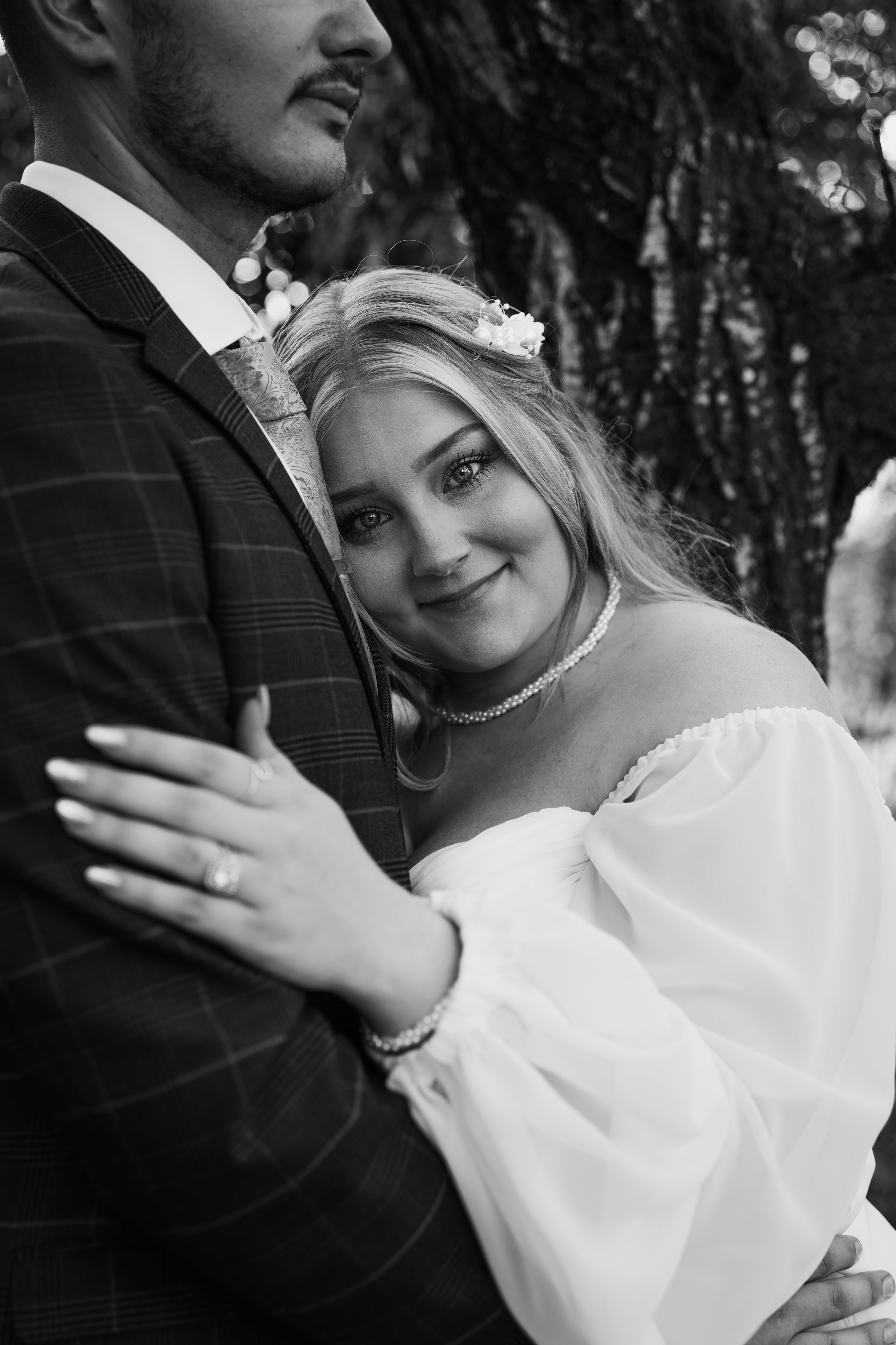 A black-and-white photo of a couple, with the woman resting her head on the man's chest, both dressed in wedding attire, outdoors near a tree.