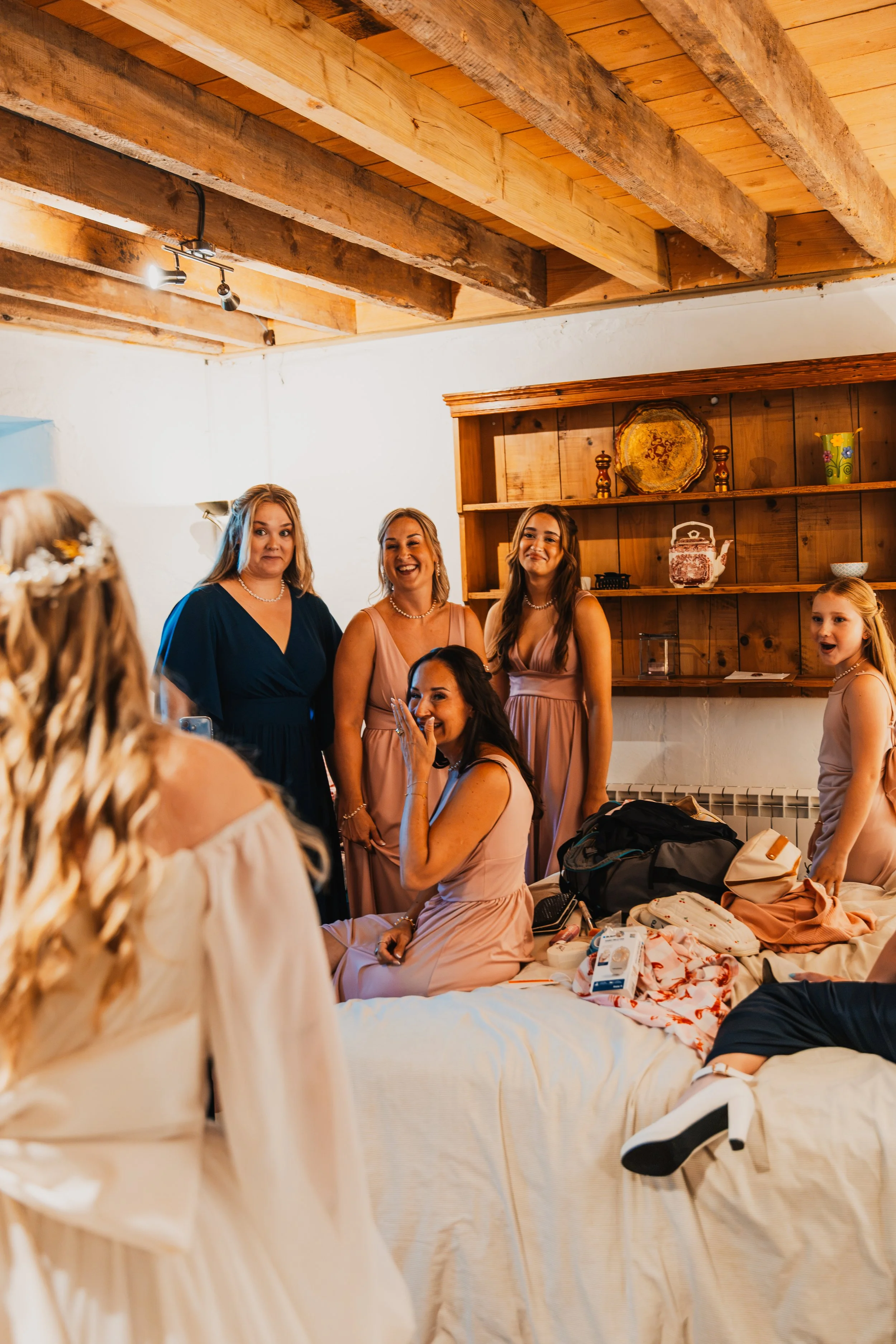 Group of women in formal dresses gathered in a cozy room with wooden ceiling beams and a wooden shelf, engaging in conversation and smiling.