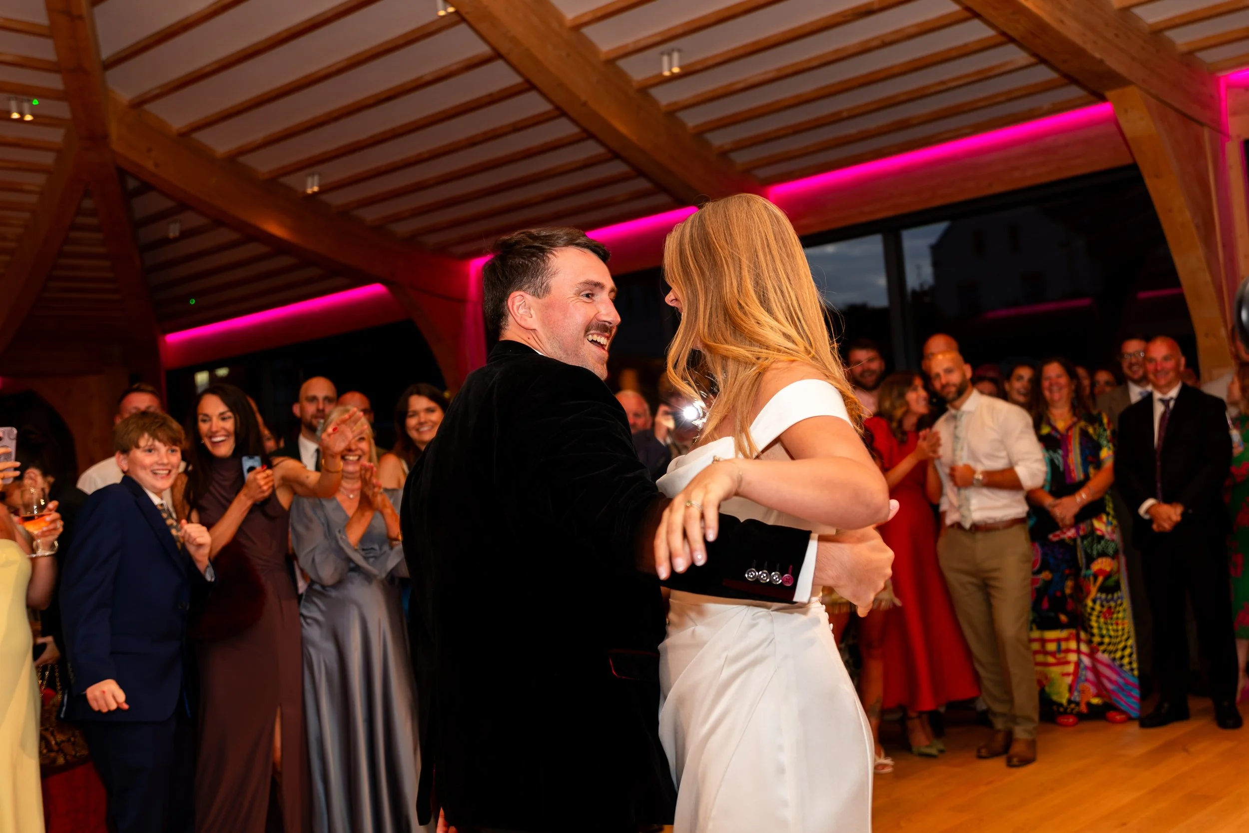 A couple dancing and smiling at a party or celebration with multiple people watching and smiling in the background. The setting has wooden beams with pink lighting accents.
