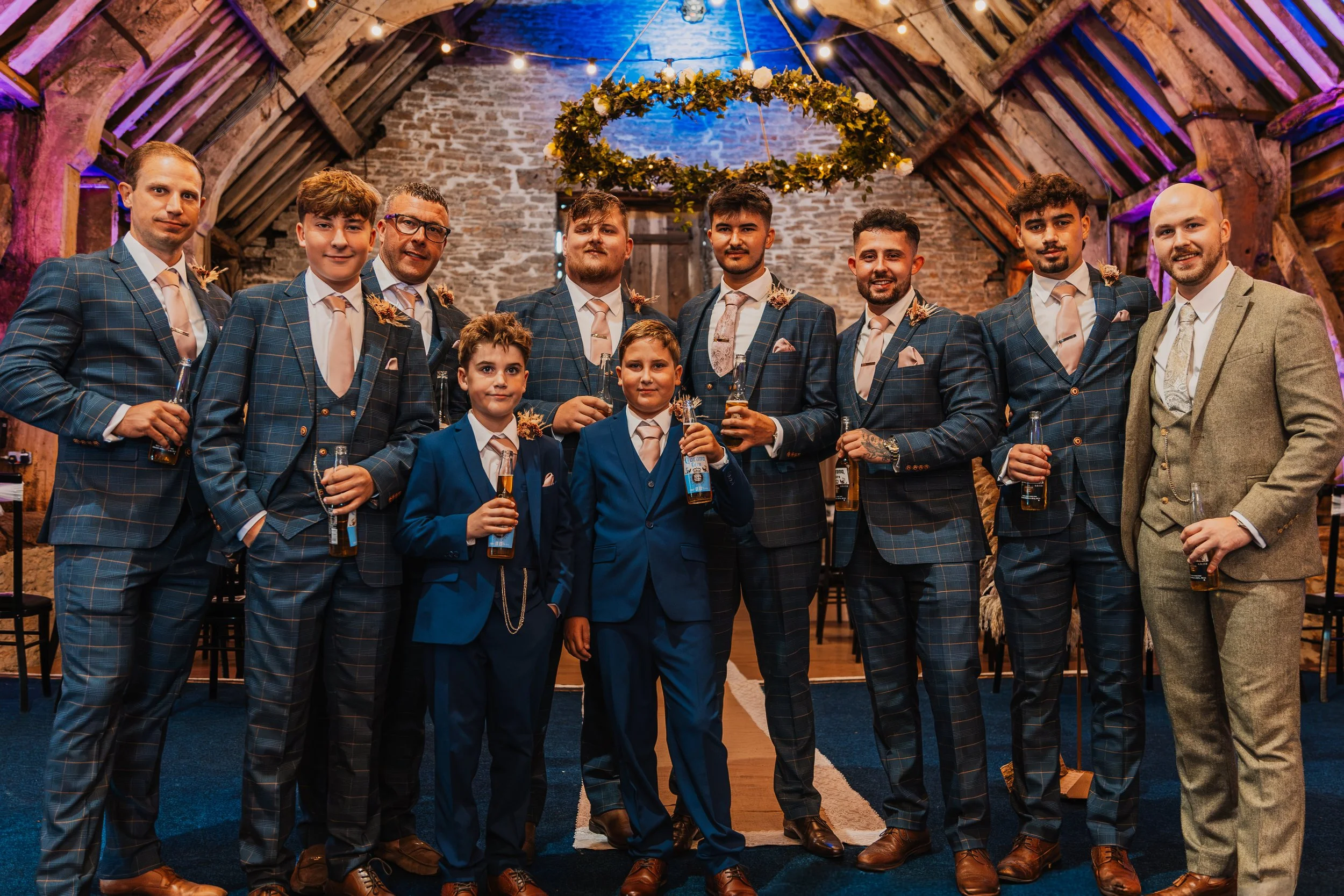 Group of men and boys dressed in formal suits, standing together inside a rustic barn with a decorative wreath hanging from the ceiling, celebrating at a wedding reception.