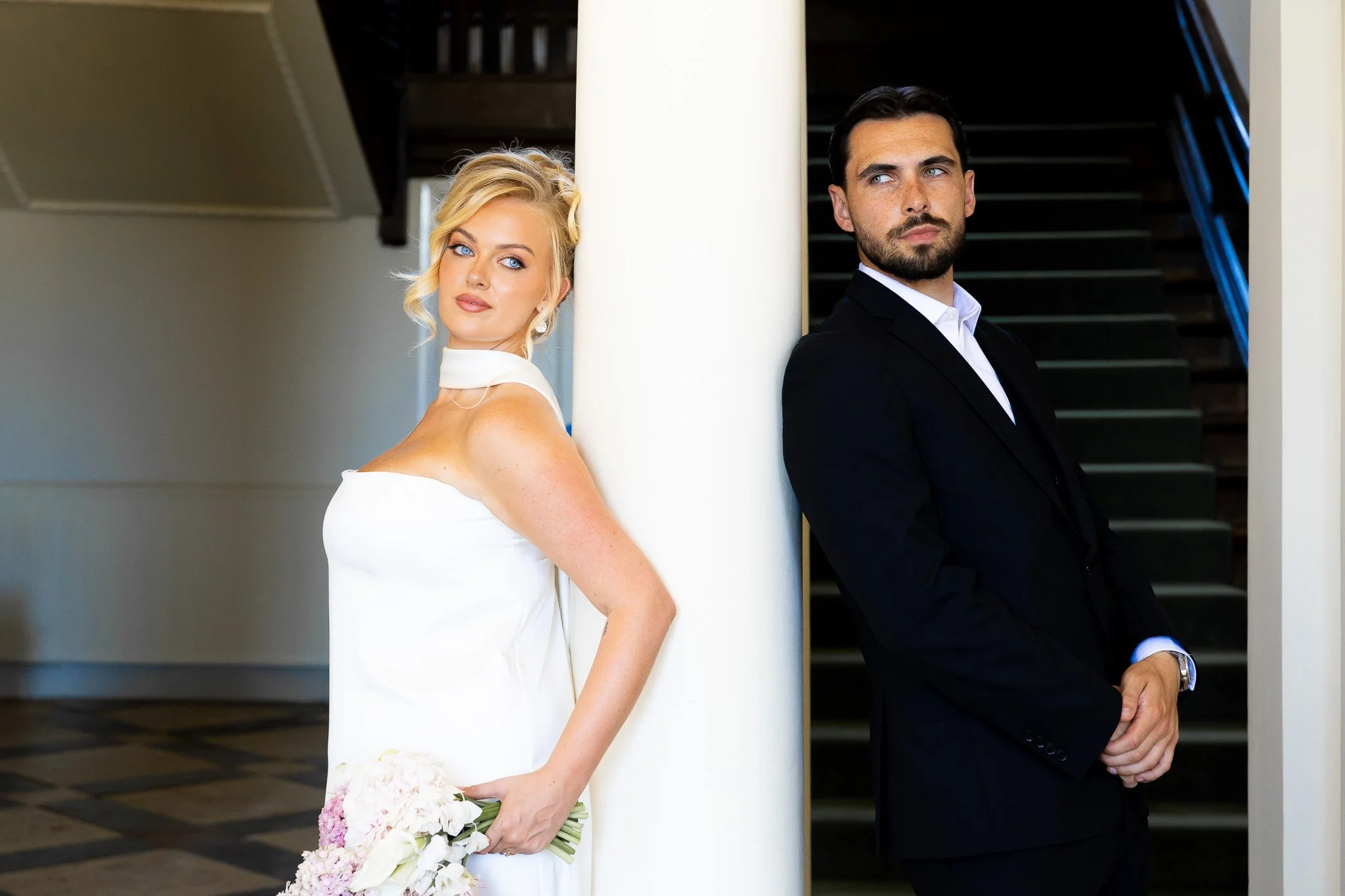 A bride and groom standing apart, back to back, separated by a white column, indoors. The bride has blonde hair styled in loose curls, wearing a white wedding dress and holding a bouquet of flowers. The groom has dark hair and a beard, wearing a blac