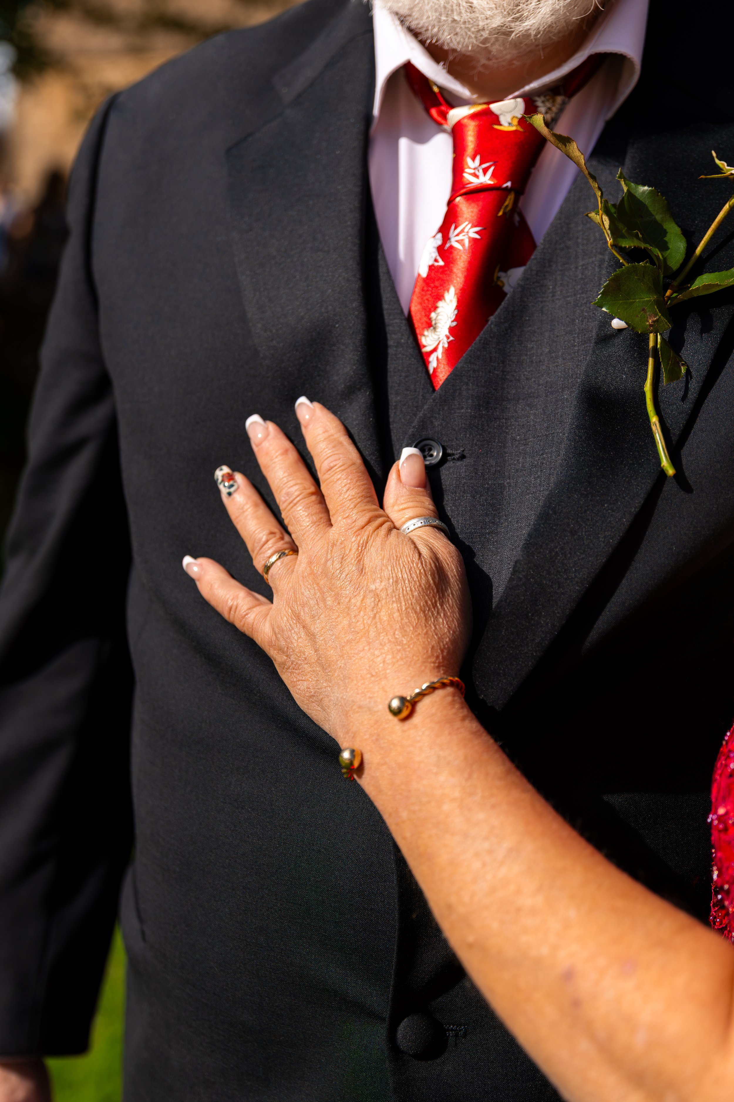 Close-up of a person wearing a black suit with a white shirt and a red floral tie. A hand with jewelry is resting on the suit, and there are green leaves attached to the suit's lapel.