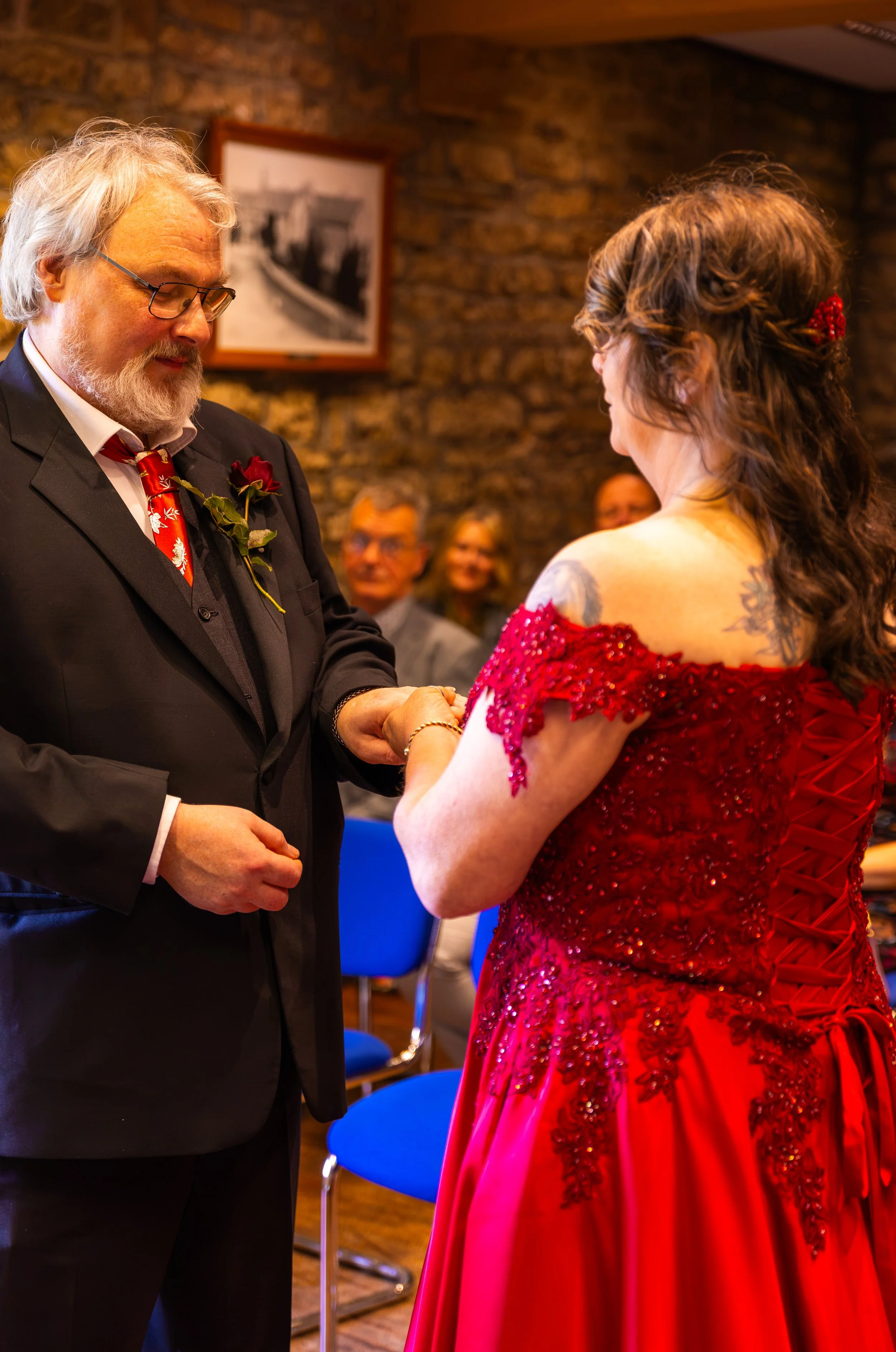 A man and woman exchange wedding vows indoors. The man has white hair, a beard, glasses, and wears a black suit with a red and white patterned tie and a red rose boutonniere. The woman has brown hair styled with a red hair accessory and wears a red d