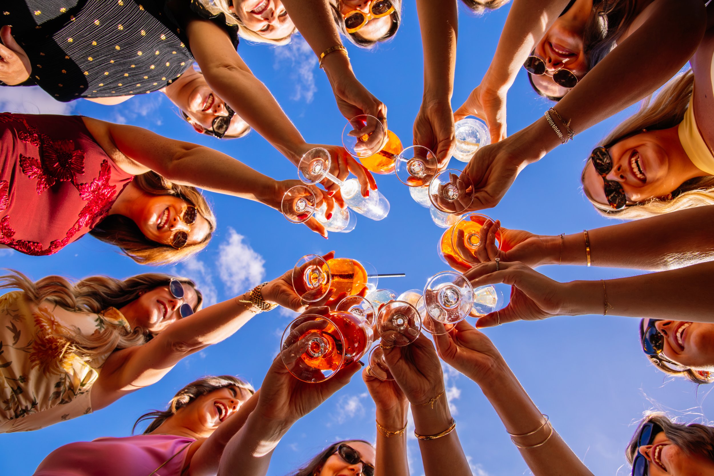 A group of women wearing sunglasses raising wine glasses together in a toast outdoors against a blue sky.
