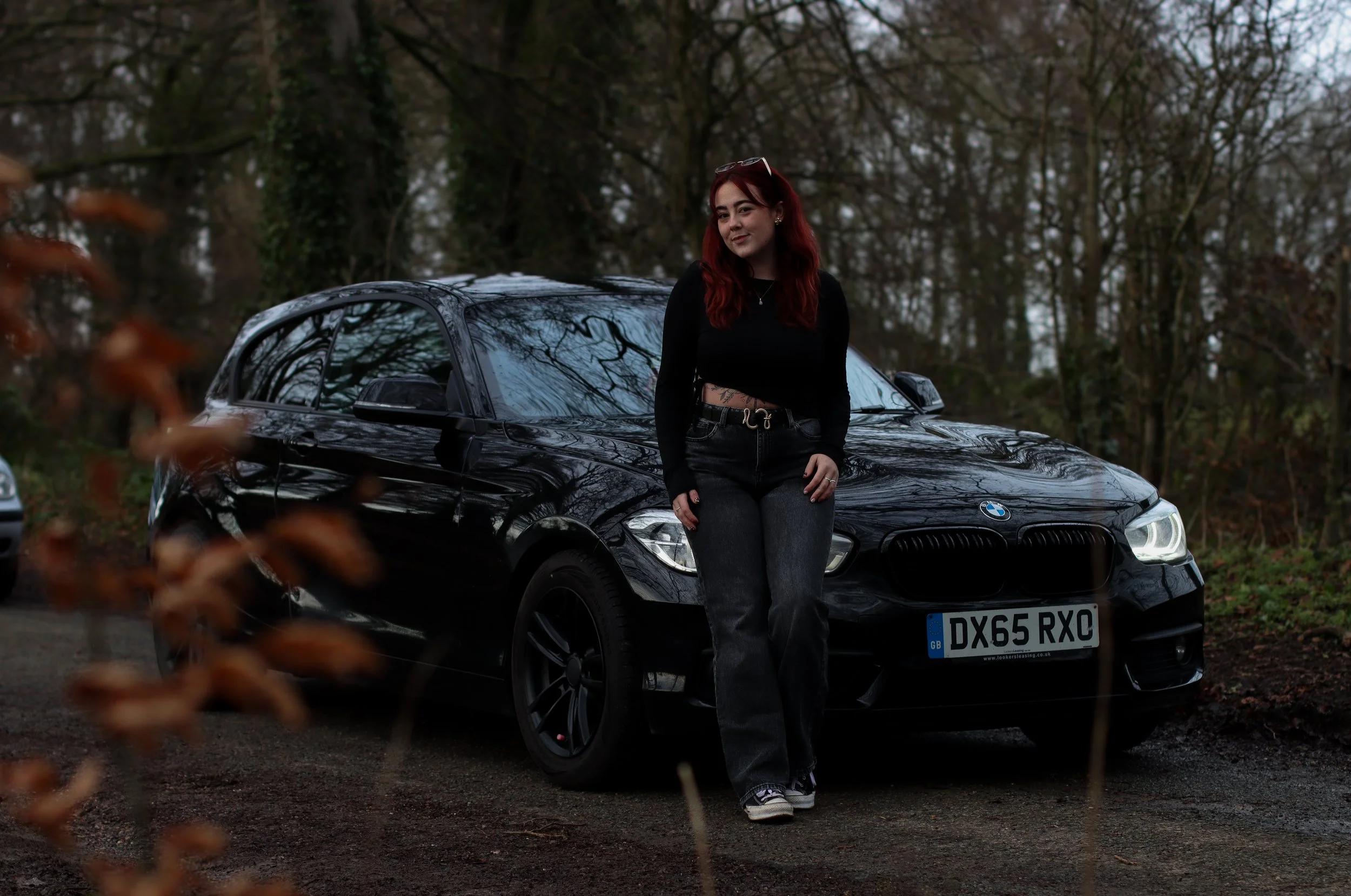 A young woman with red hair and a black outfit leaning against a black BMW car in a wooded area during overcast weather.