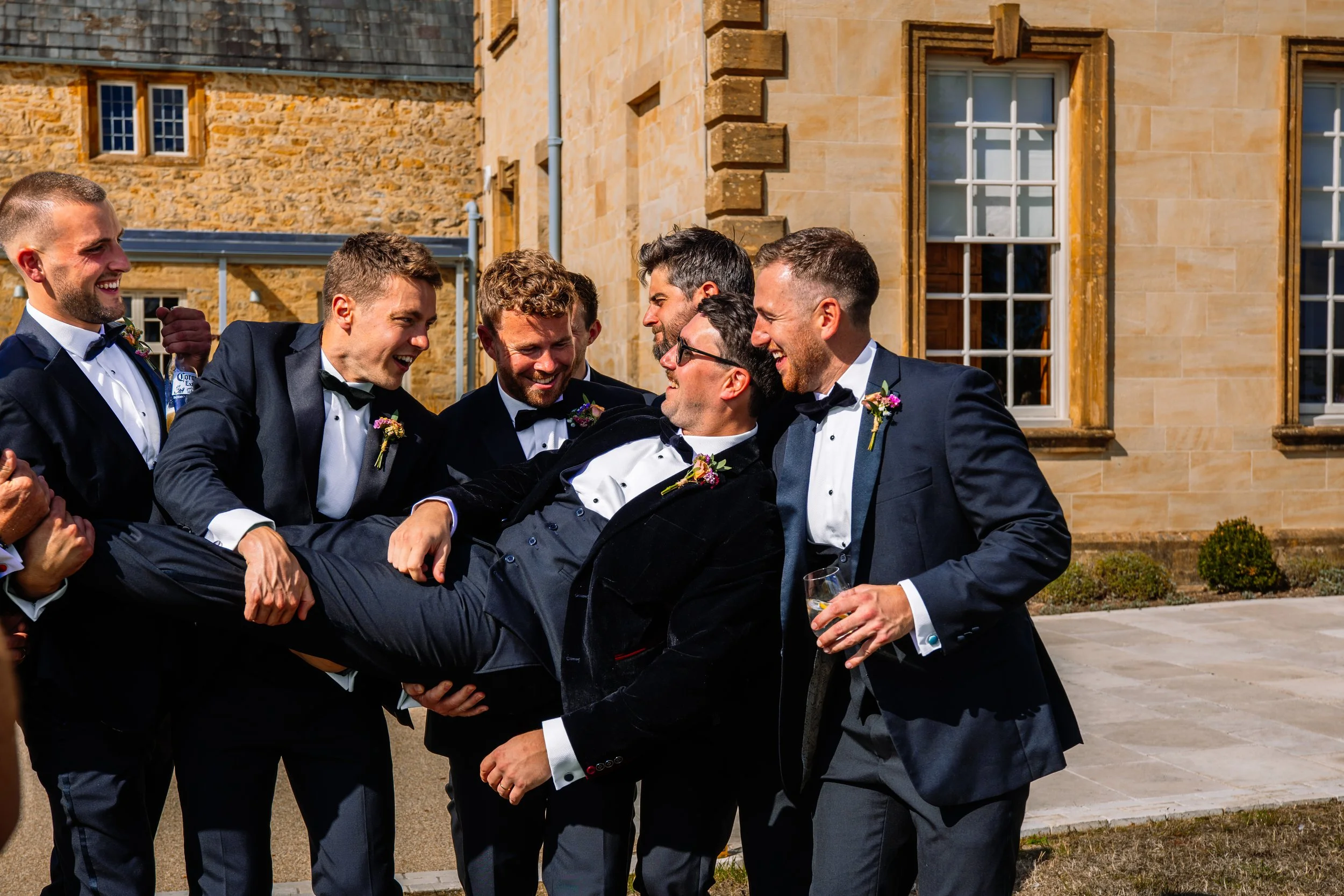 Group of men dressed in tuxedos, some with boutonnières, joyfully carrying a man in a tuxedo with a microphone outside a stone building during daytime.