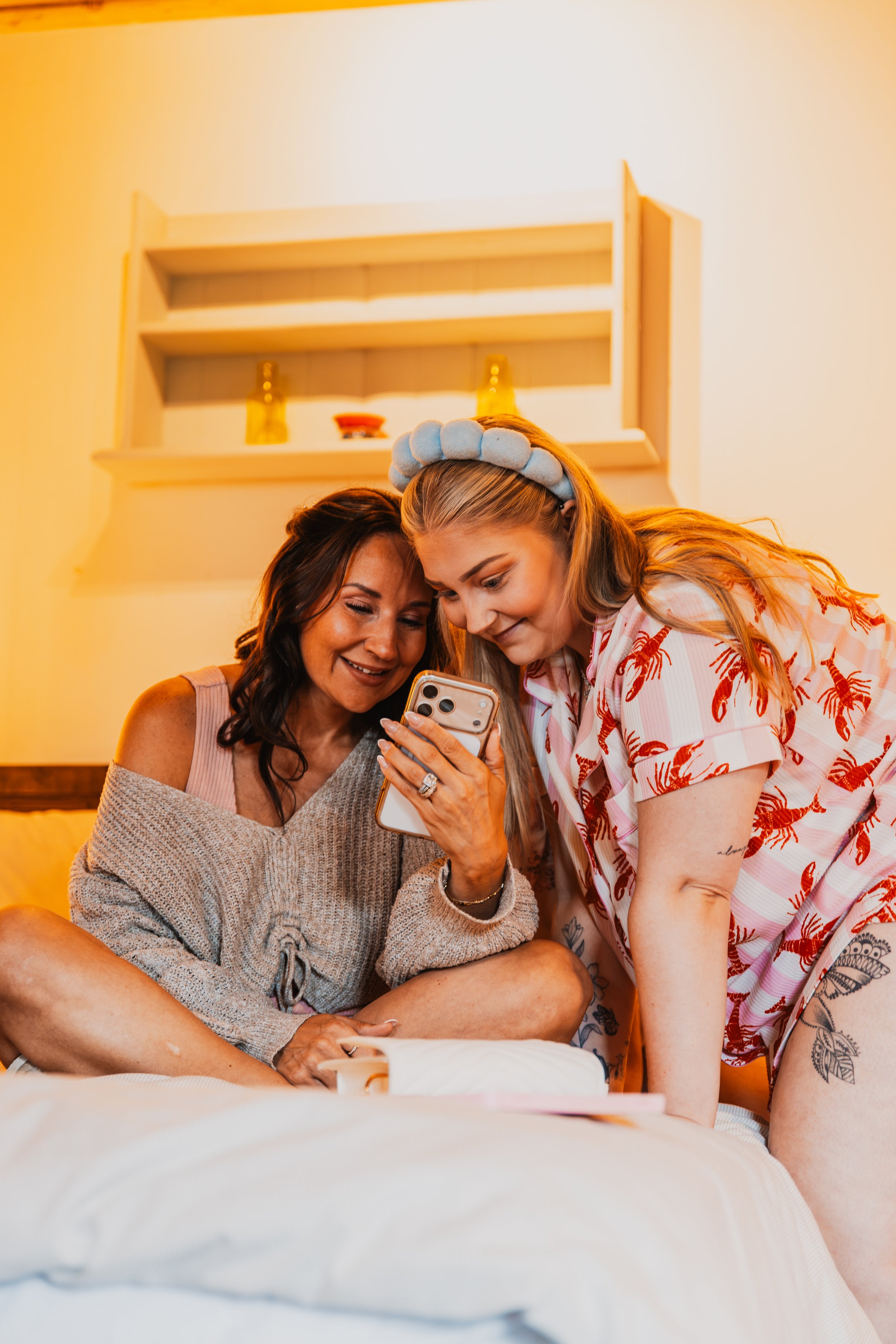 Two women, one with dark hair and the other blonde wearing a headband, are looking at a phone and smiling while sitting on a bed with white bedding. A wooden shelf with decorations is on the wall behind them.