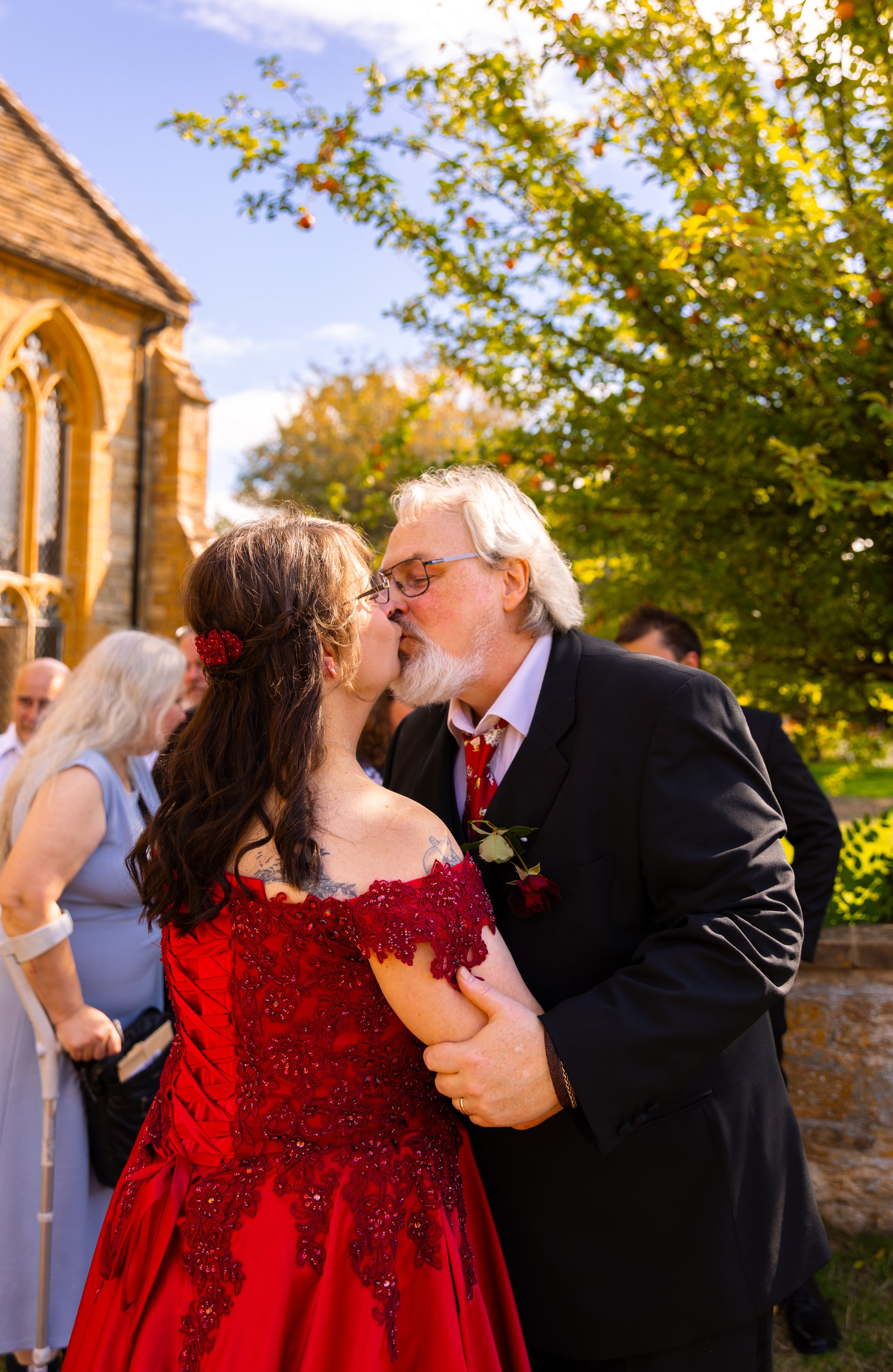 A couple is kissing during a wedding ceremony outdoors, with a woman in a lavender dress and other guests in the background, next to a building and a tree with green leaves and some orange fruits.