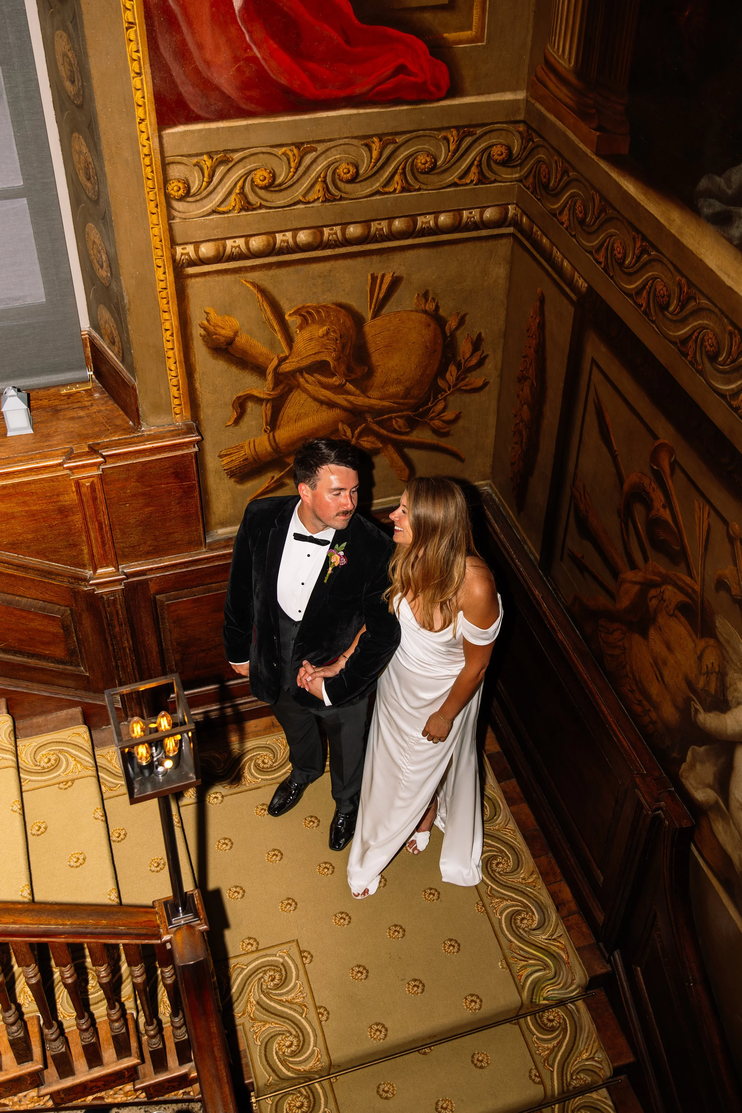 A man and woman in formal attire standing on a staircase, looking at each other and holding hands. The man is wearing a black tuxedo with a bow tie and boutonniere, and the woman is wearing a strapless white gown. The staircase has ornate wooden pane