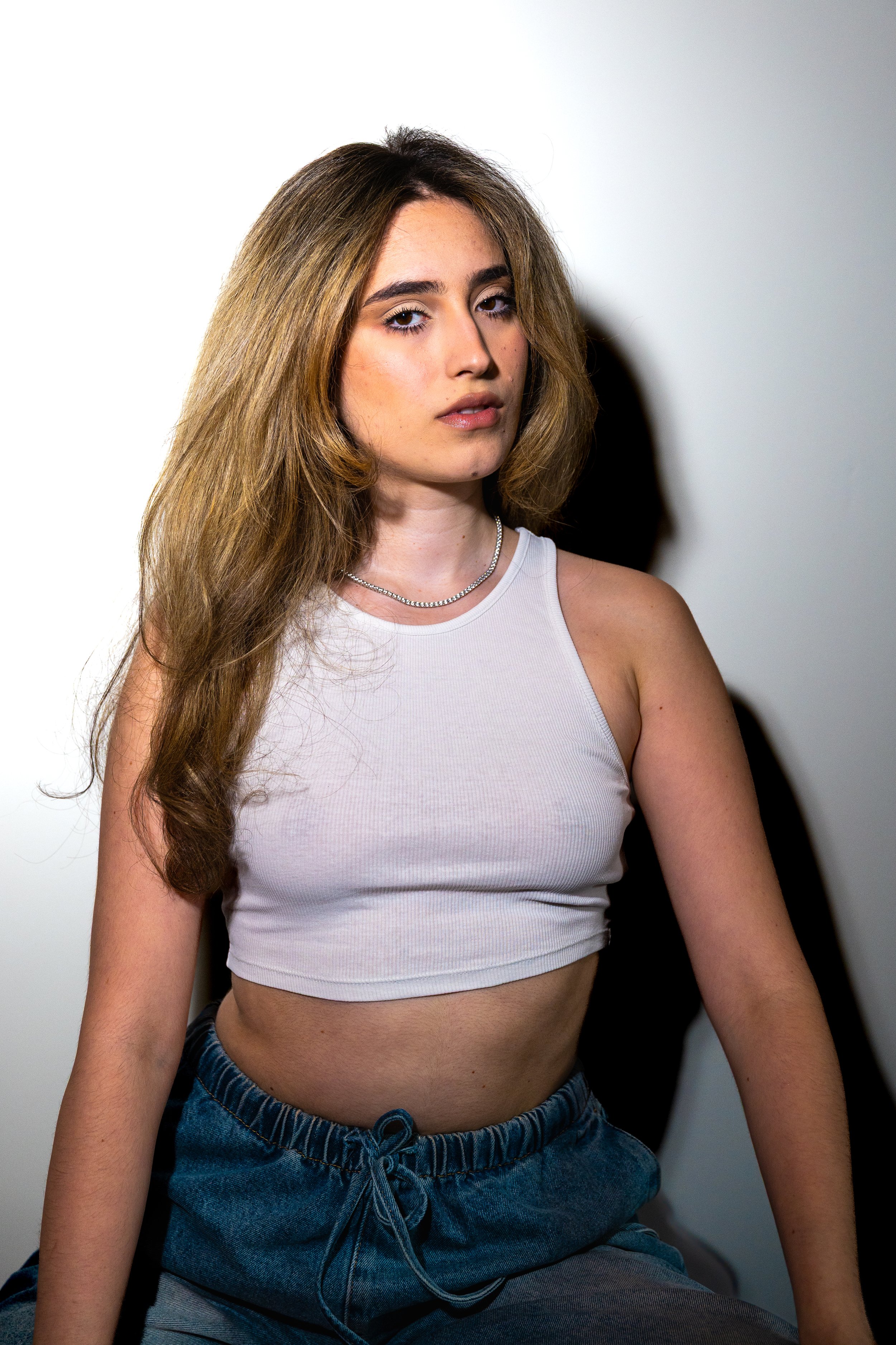 A young woman with long, wavy brown hair, wearing a white tank top, blue denim shorts with a drawstring, and a silver necklace, sitting against a white background.
