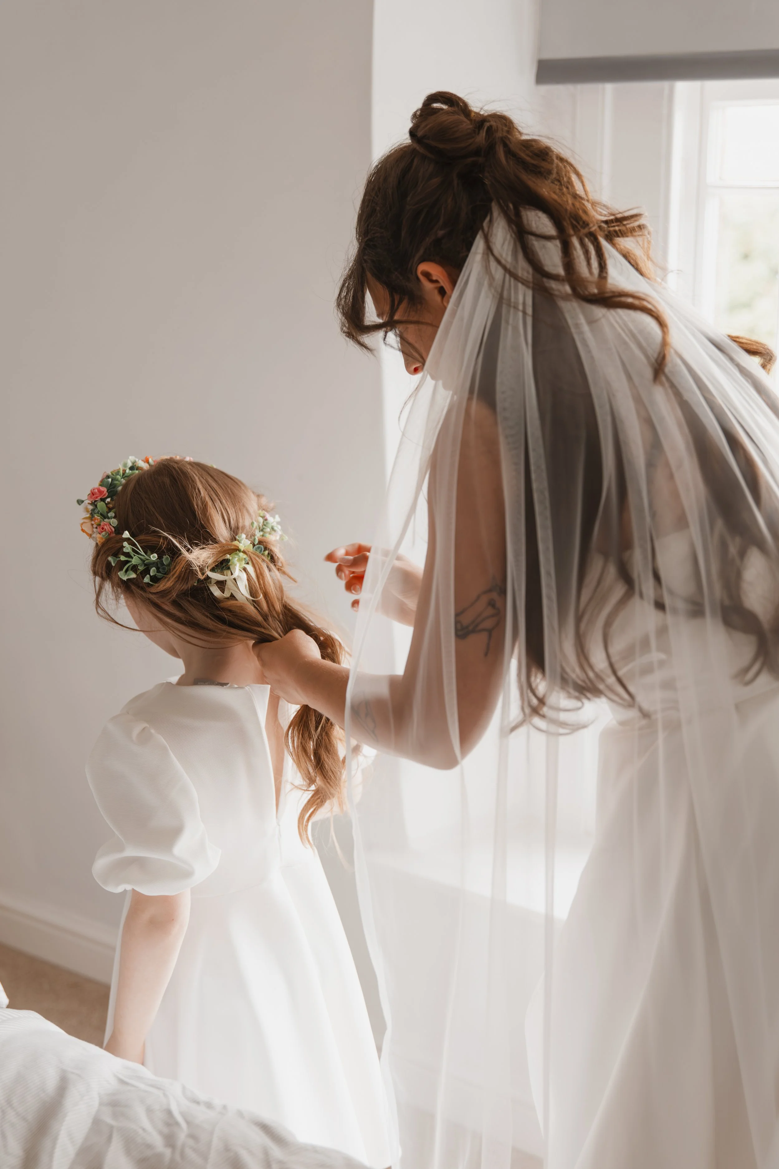 A woman dressed in a white wedding gown is adjusting the dress of a young girl who is also in a white dress with puffed sleeves, wearing a floral crown, as they stand near a window with sunlight streaming in.