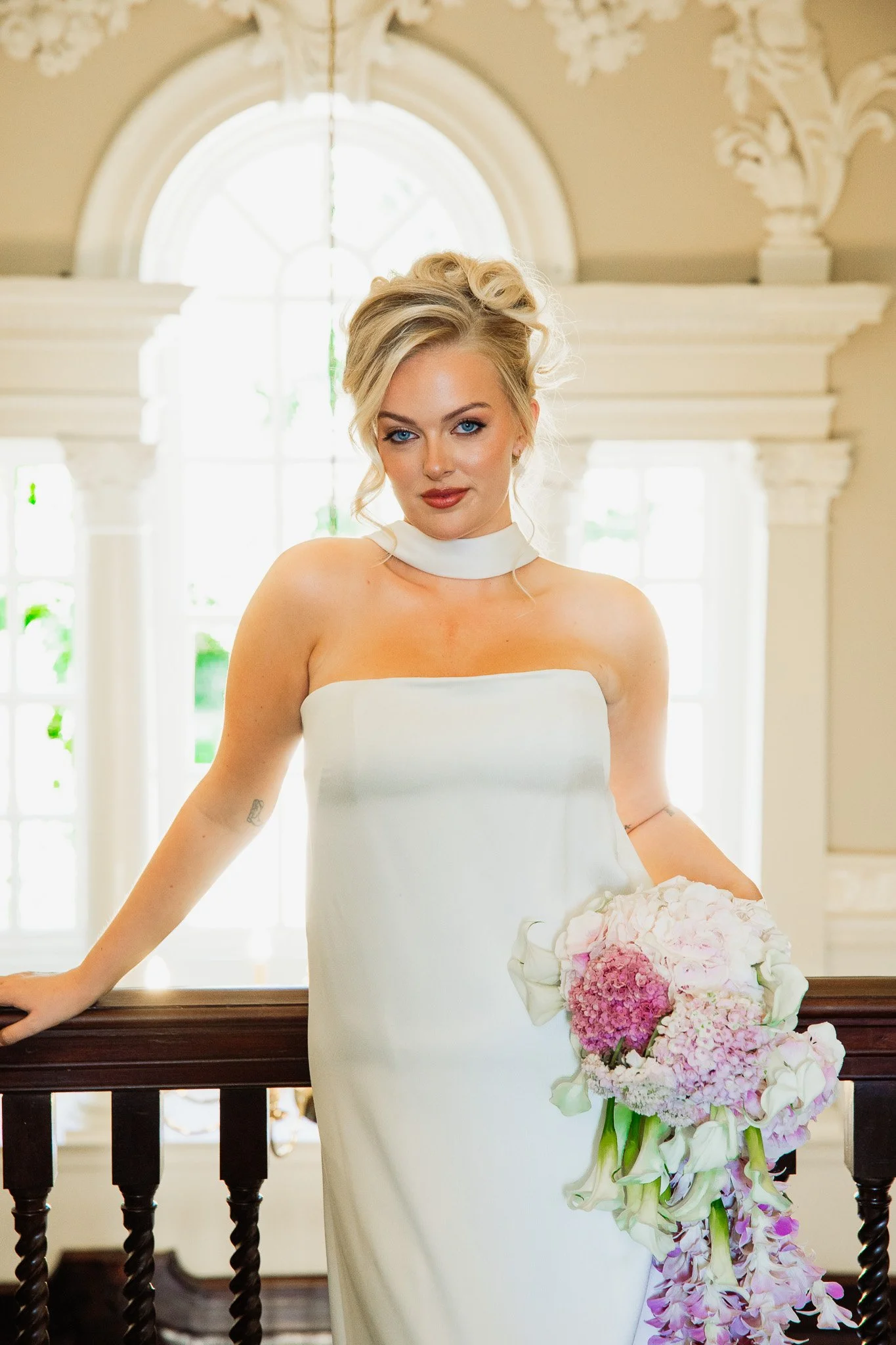 A woman in a strapless white wedding gown holding a cascading bouquet of pink and white flowers, standing inside a bright, ornate room with large arched windows in the background.