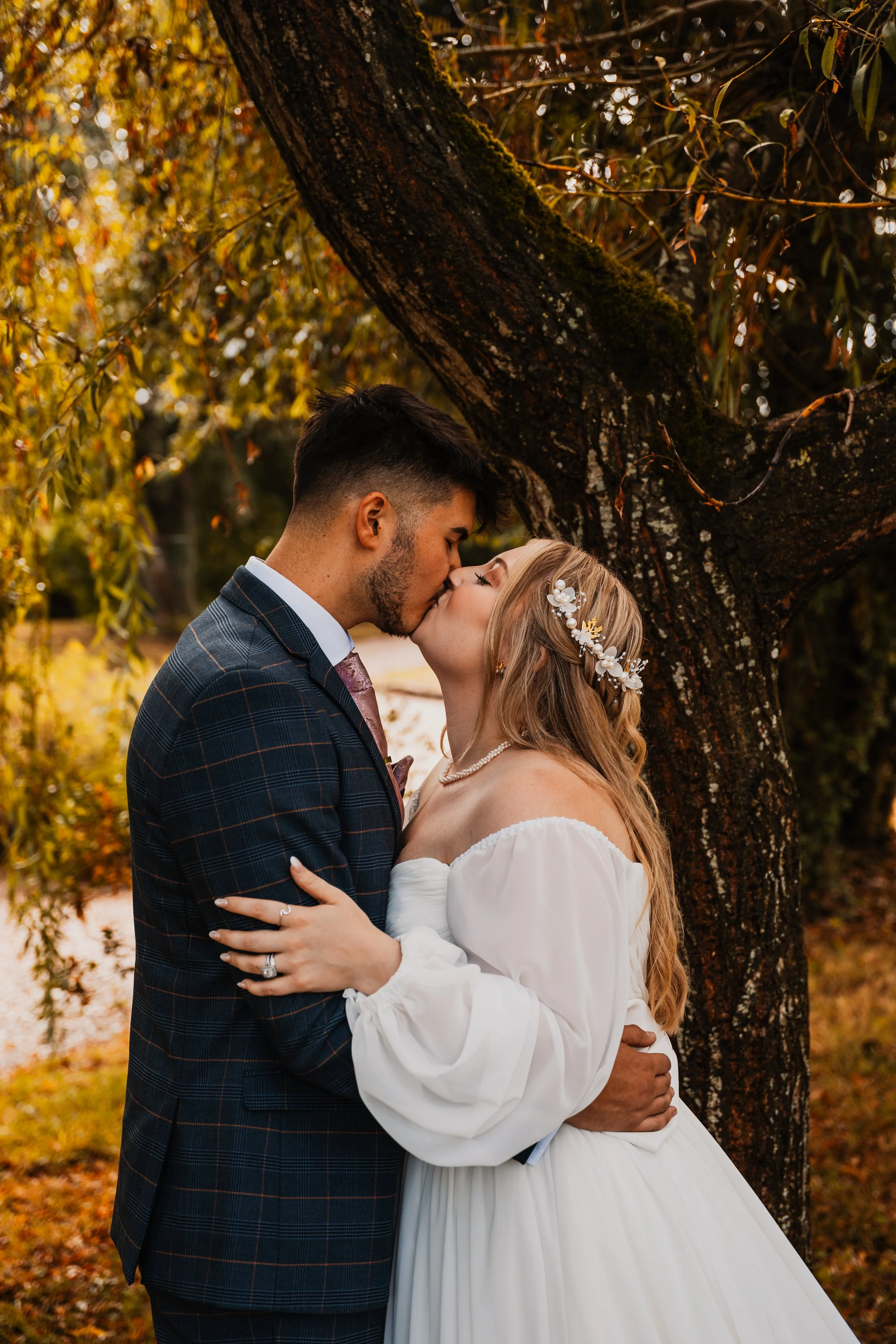 A couple sharing a kiss outdoors during autumn, with trees and leaves in fall colors in the background.