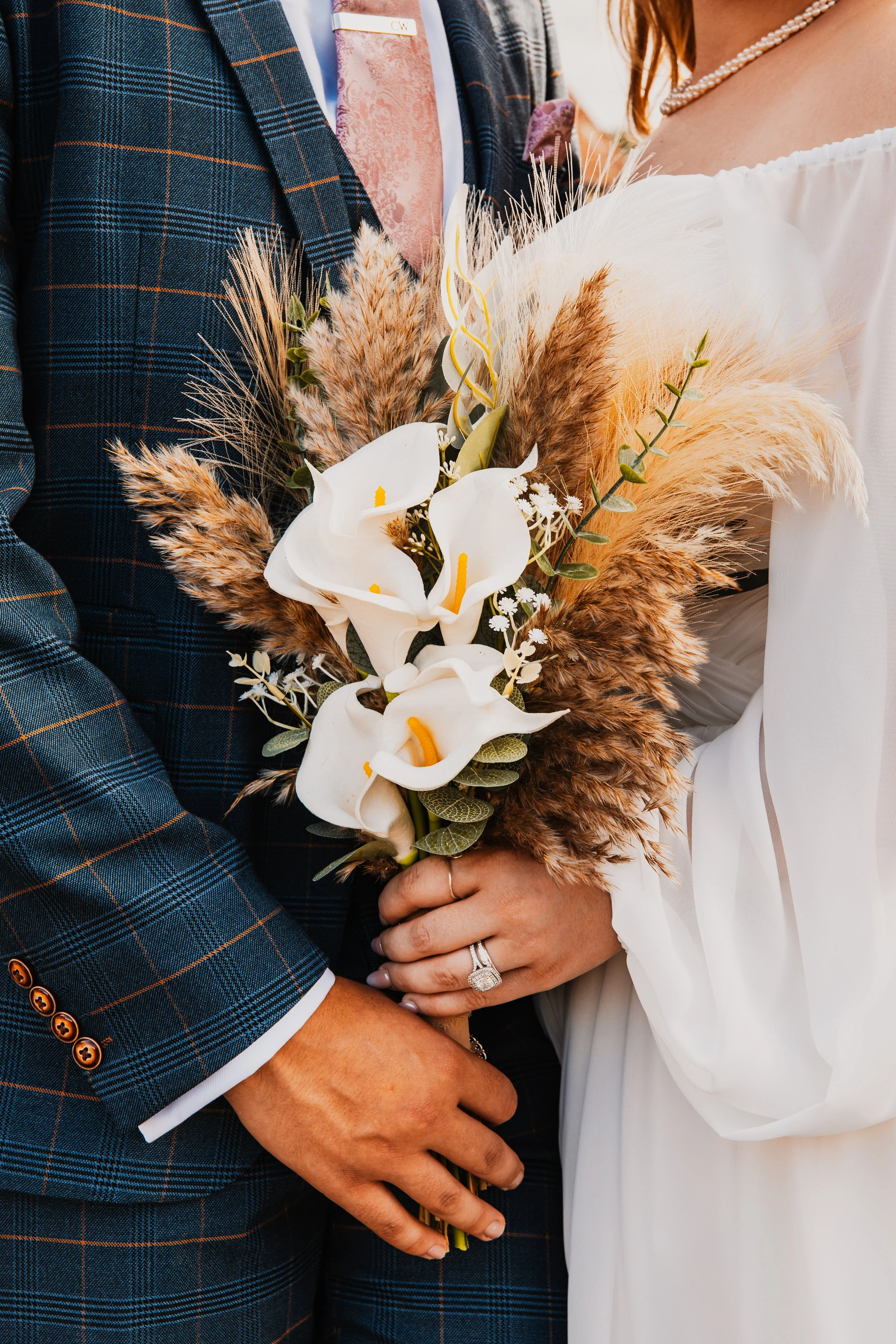 Close-up of a couple at their wedding, with the bride holding a bouquet of white calla lilies, pampas grass, and greenery, as they embrace.